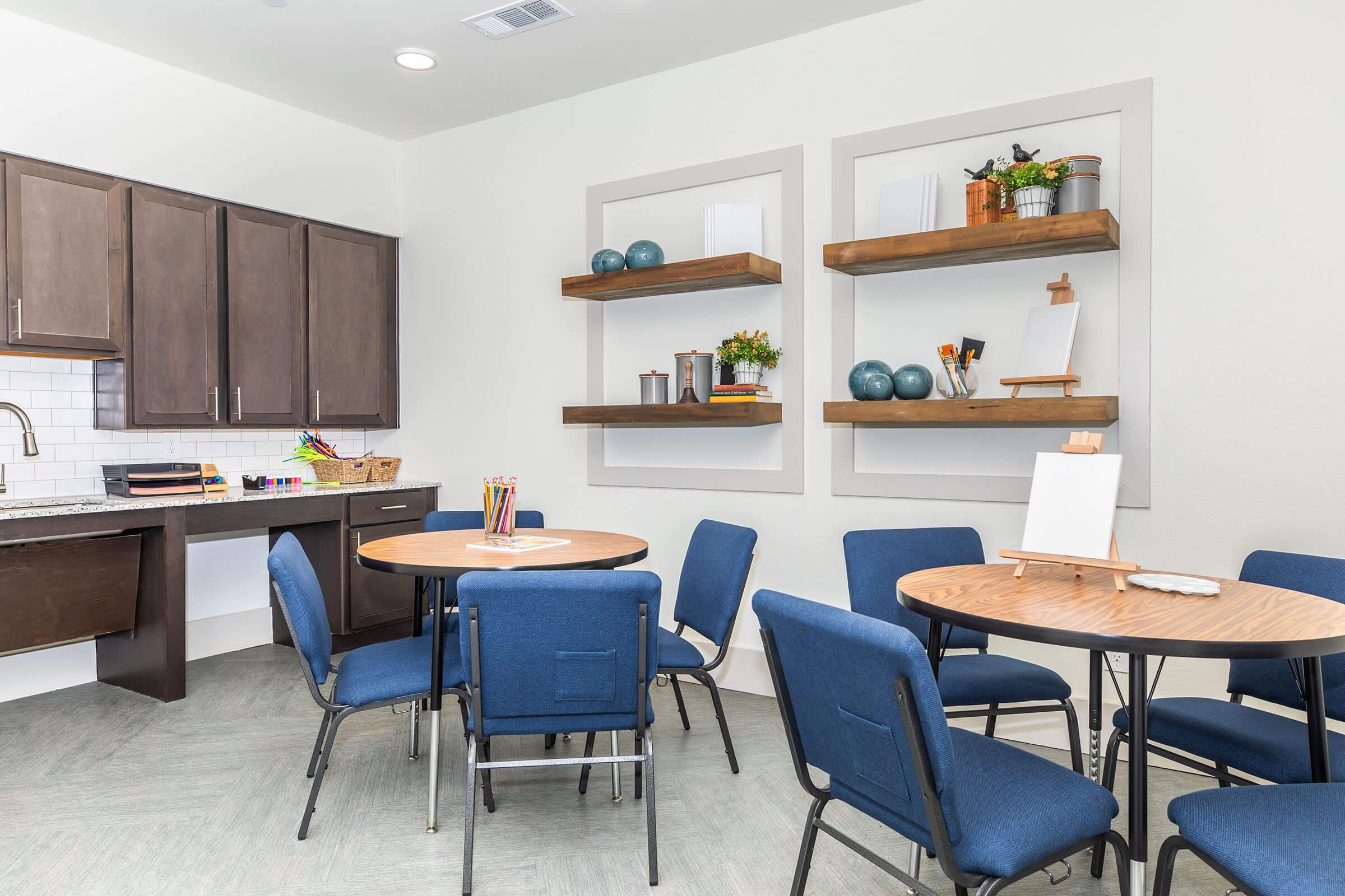 A bright room featuring two round tables surrounded by blue chairs. One table has art supplies and an easel, while the other is clear. The walls display decorative shelves with plants and objects, and there is a kitchen area with dark cabinetry and a countertop in the background.