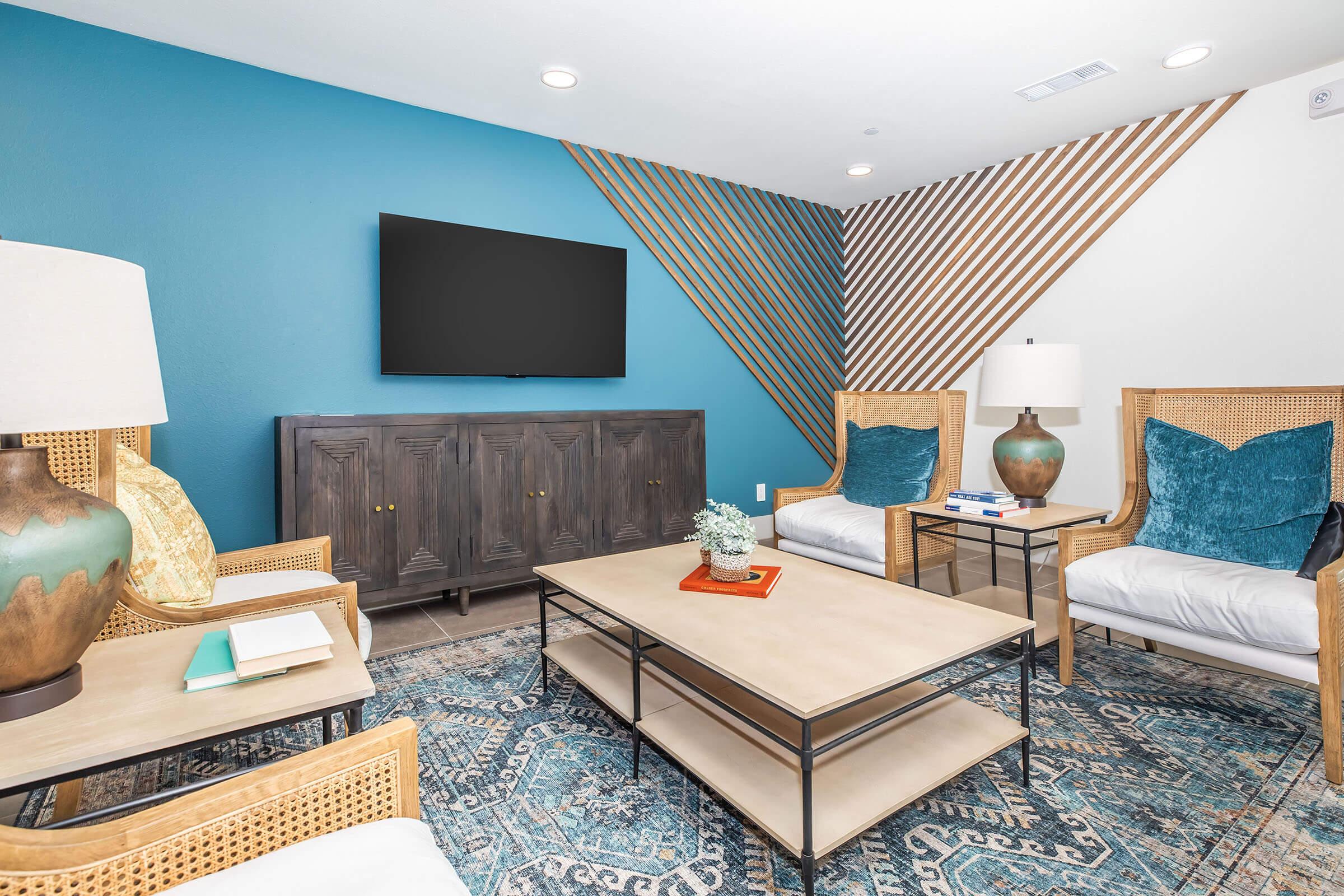 A cozy living room featuring two white cushioned chairs with blue throw pillows, a coffee table with a decorative item, and a modern entertainment center. The walls are painted in a striking blue, complemented by a unique wooden design. A large flat-screen TV is mounted on the wall, and a stylish lamp adds warmth to the space.