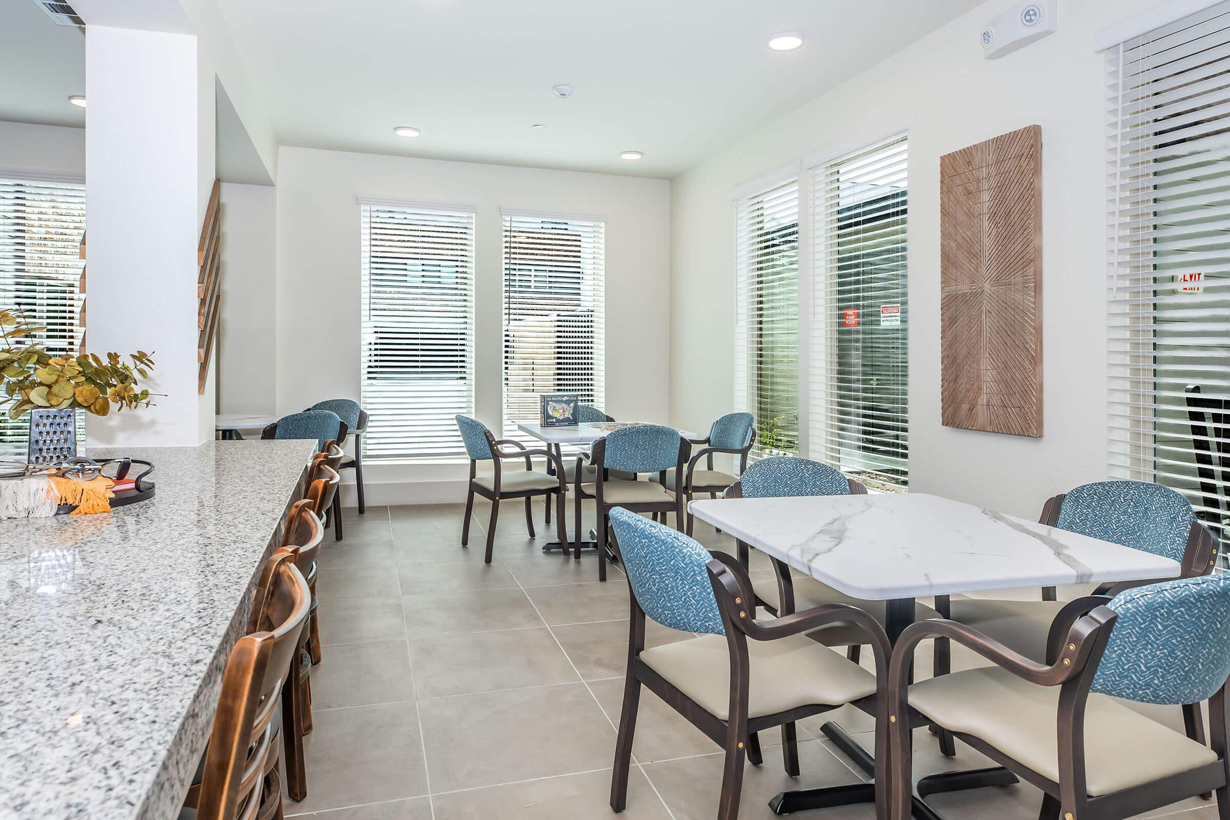 Bright and modern dining area featuring a marble-topped table surrounded by patterned chairs. The space includes large windows with blinds, allowing natural light to fill the room. A granite countertop bar is visible on one side, enhancing the contemporary feel of the interior.