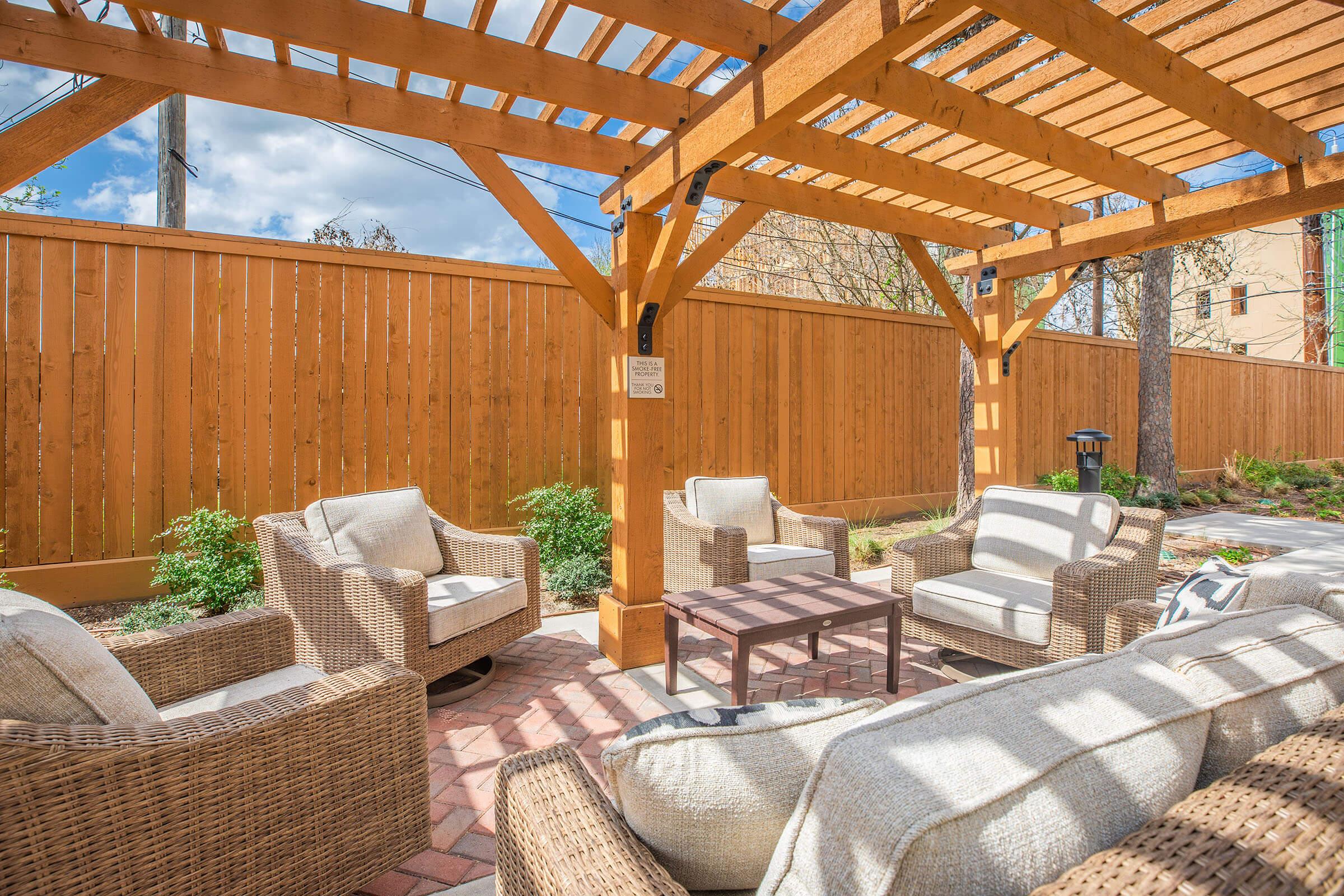 A cozy outdoor seating area under a wooden pergola, featuring four cushioned chairs arranged around a small table. The space is decorated with greenery and has a wooden fence in the background, with blue skies and clouds visible above.