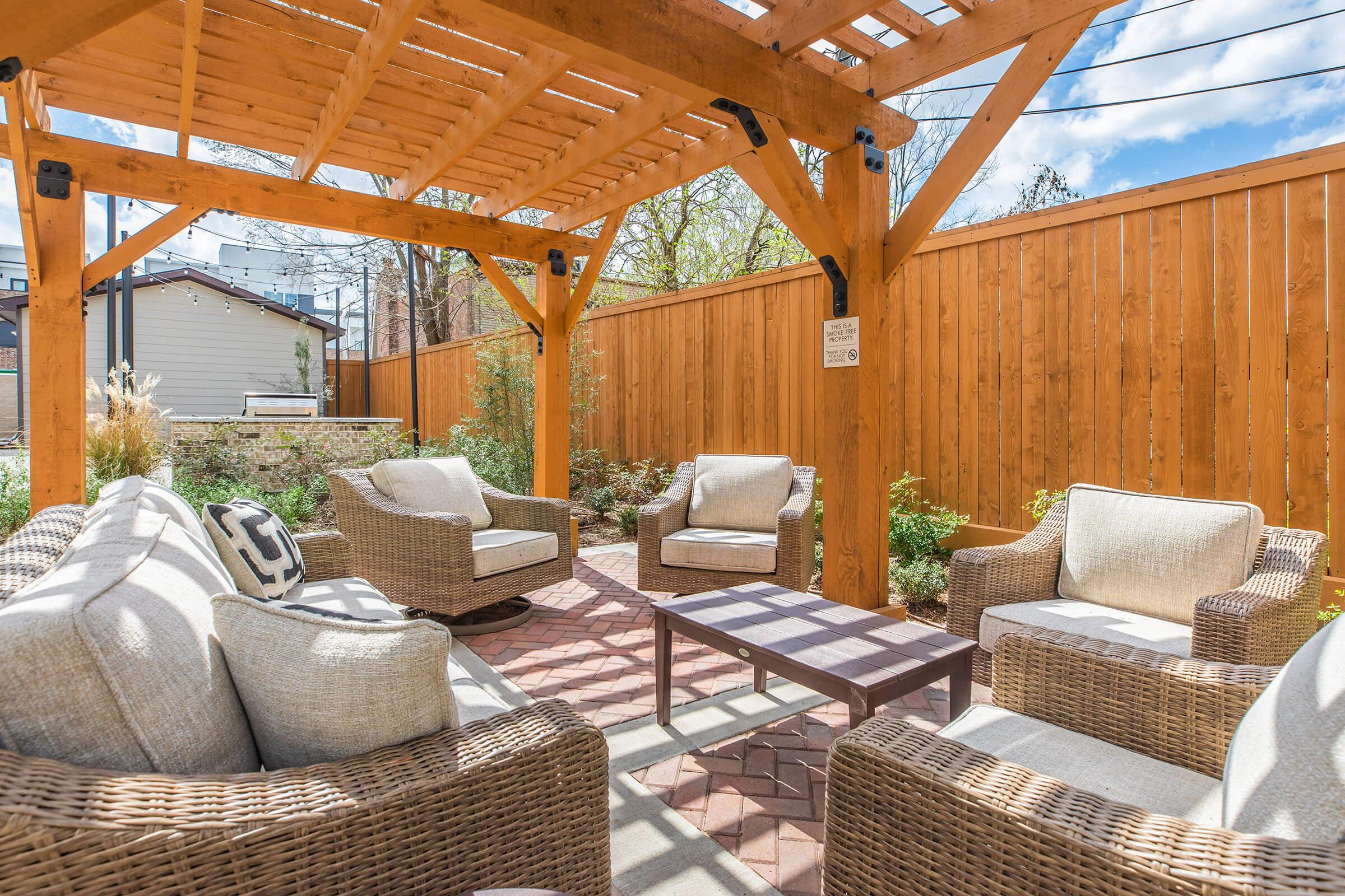 A cozy outdoor seating area featuring four wicker chairs and a small table, situated under a wooden pergola. Surrounding greenery and a wooden fence create a serene atmosphere, with blue skies and clouds visible above. Perfect for relaxation and gatherings.