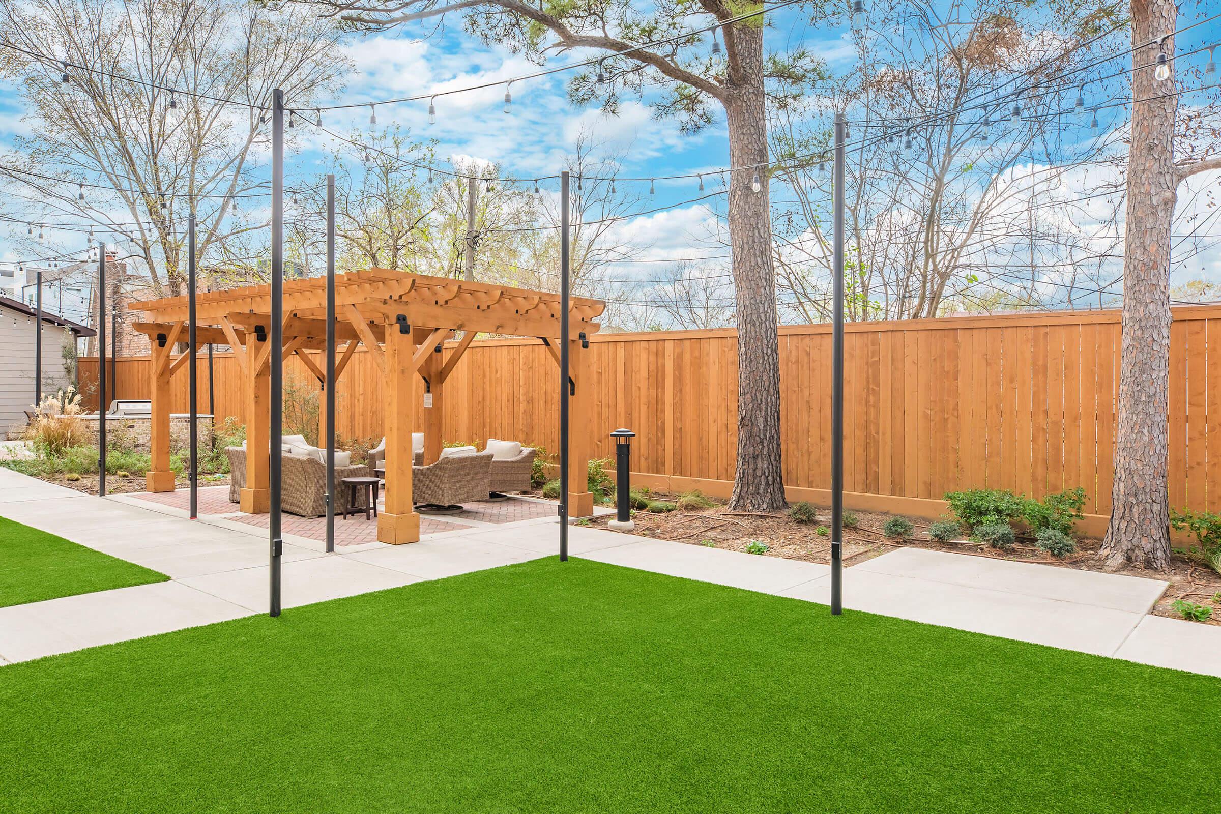 A landscaped backyard featuring a pergola with outdoor seating, surrounded by greenery. The area is divided by a grassy patch and concrete pathways, with wooden fencing and trees in the background, creating a serene outdoor space.