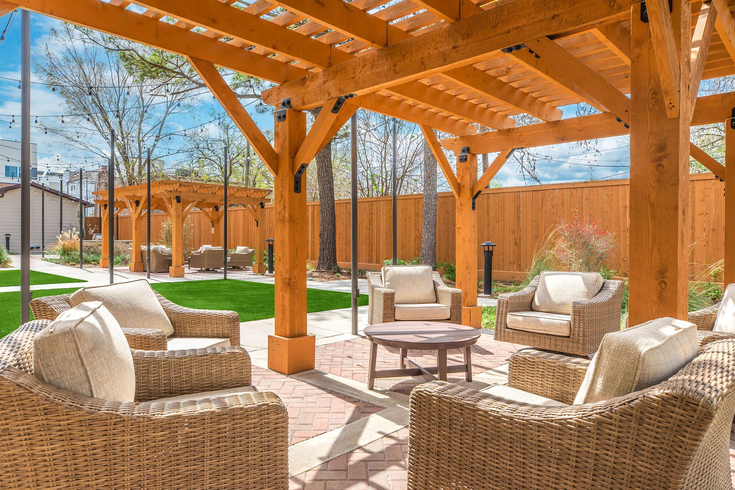 A cozy outdoor seating area featuring wicker chairs around a circular table, shaded by a wooden pergola. Lush green grass and trees provide a natural backdrop, while another pergola is visible in the background, creating a relaxing atmosphere for gatherings. Bright blue skies add to the inviting ambiance.