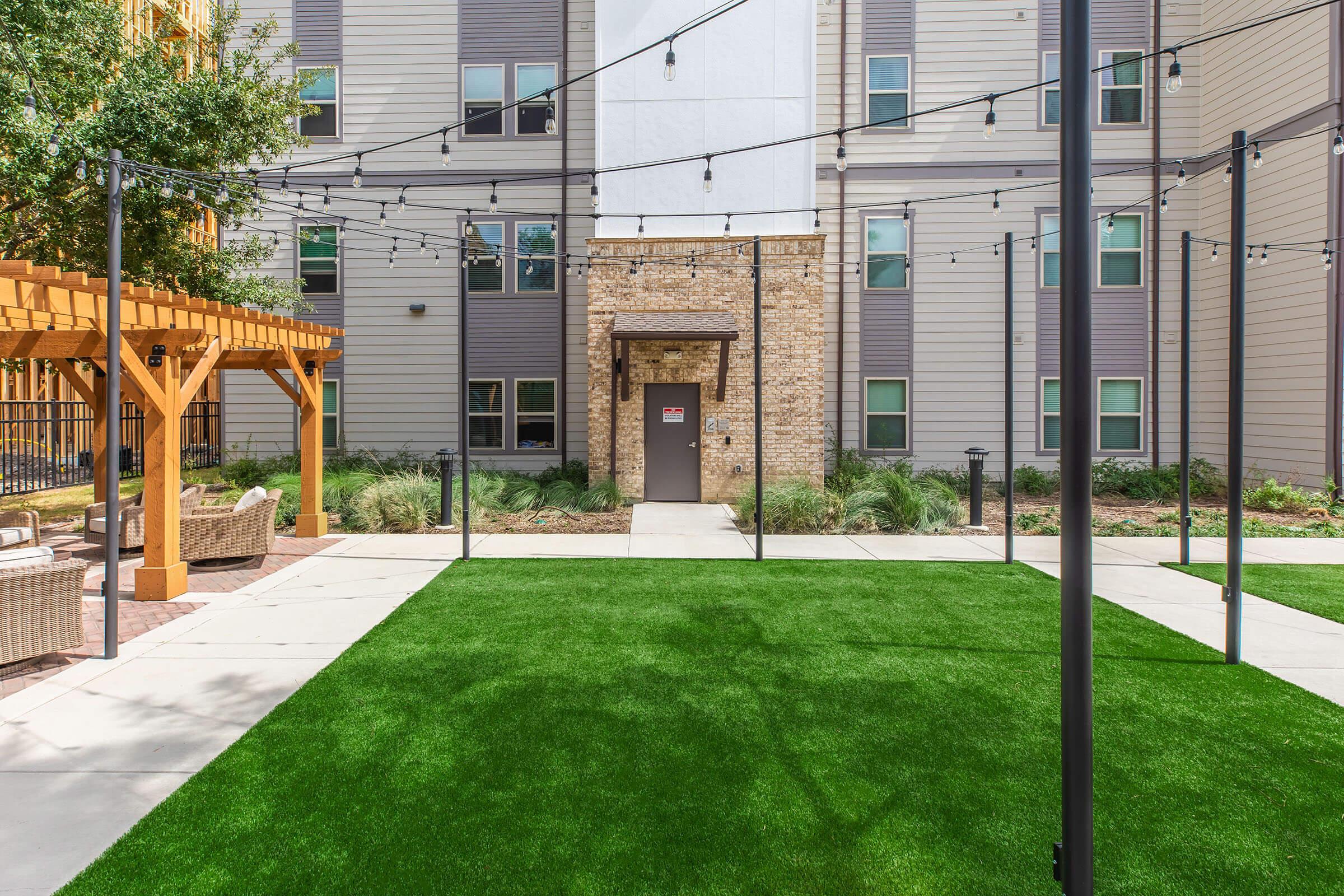 A well-maintained outdoor space featuring green artificial grass, string lights overhead, and a building entrance with stone accents. There are wooden structures and comfortable seating areas, creating an inviting atmosphere for relaxation and social gatherings in a residential setting.