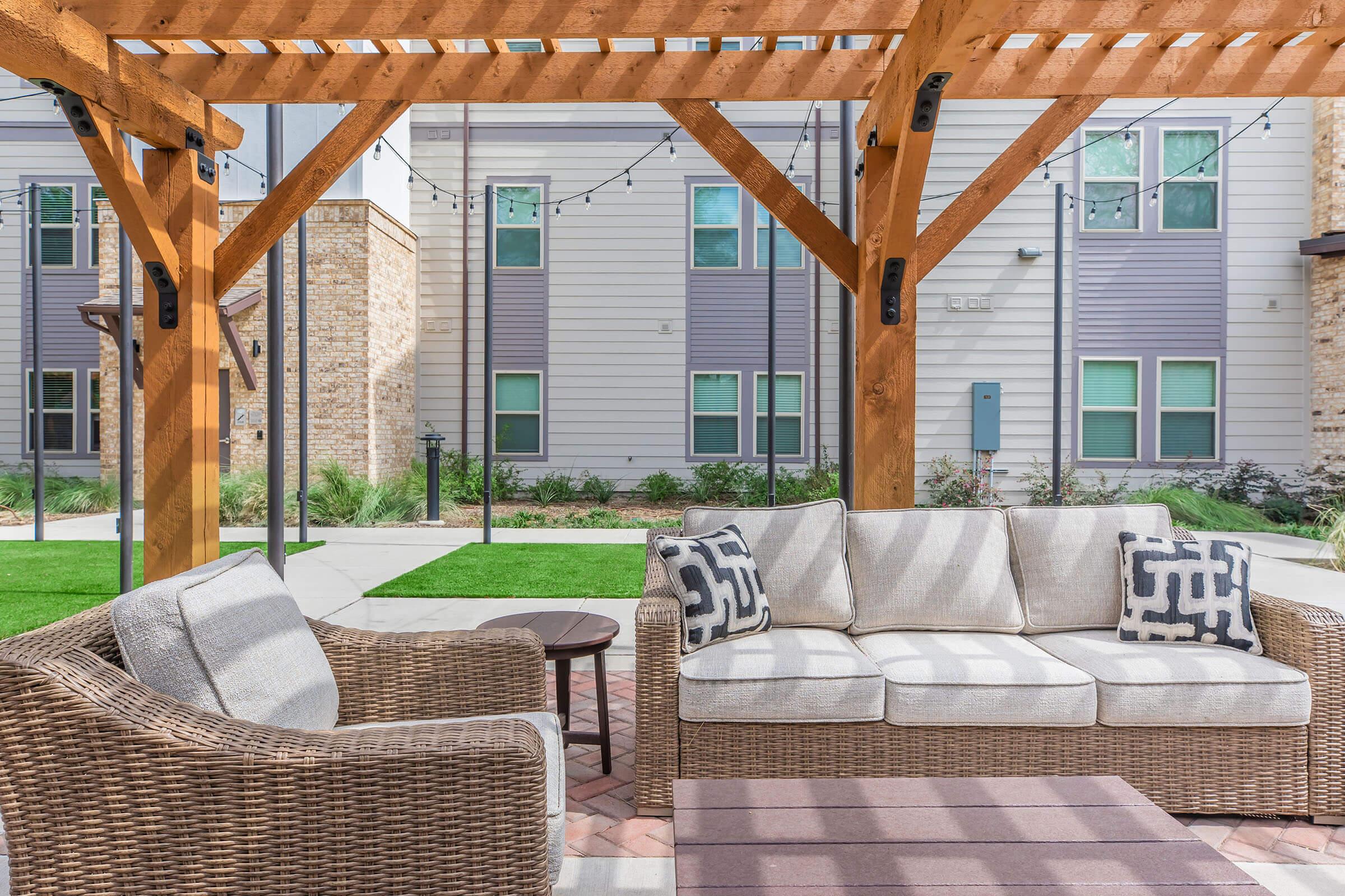 Stylish outdoor seating area with a light-colored sofa and armchair, complemented by decorative pillows. A small round table sits between the chairs. The space is shaded by a wooden pergola, with green grass and buildings in the background, creating a welcoming and comfortable atmosphere.