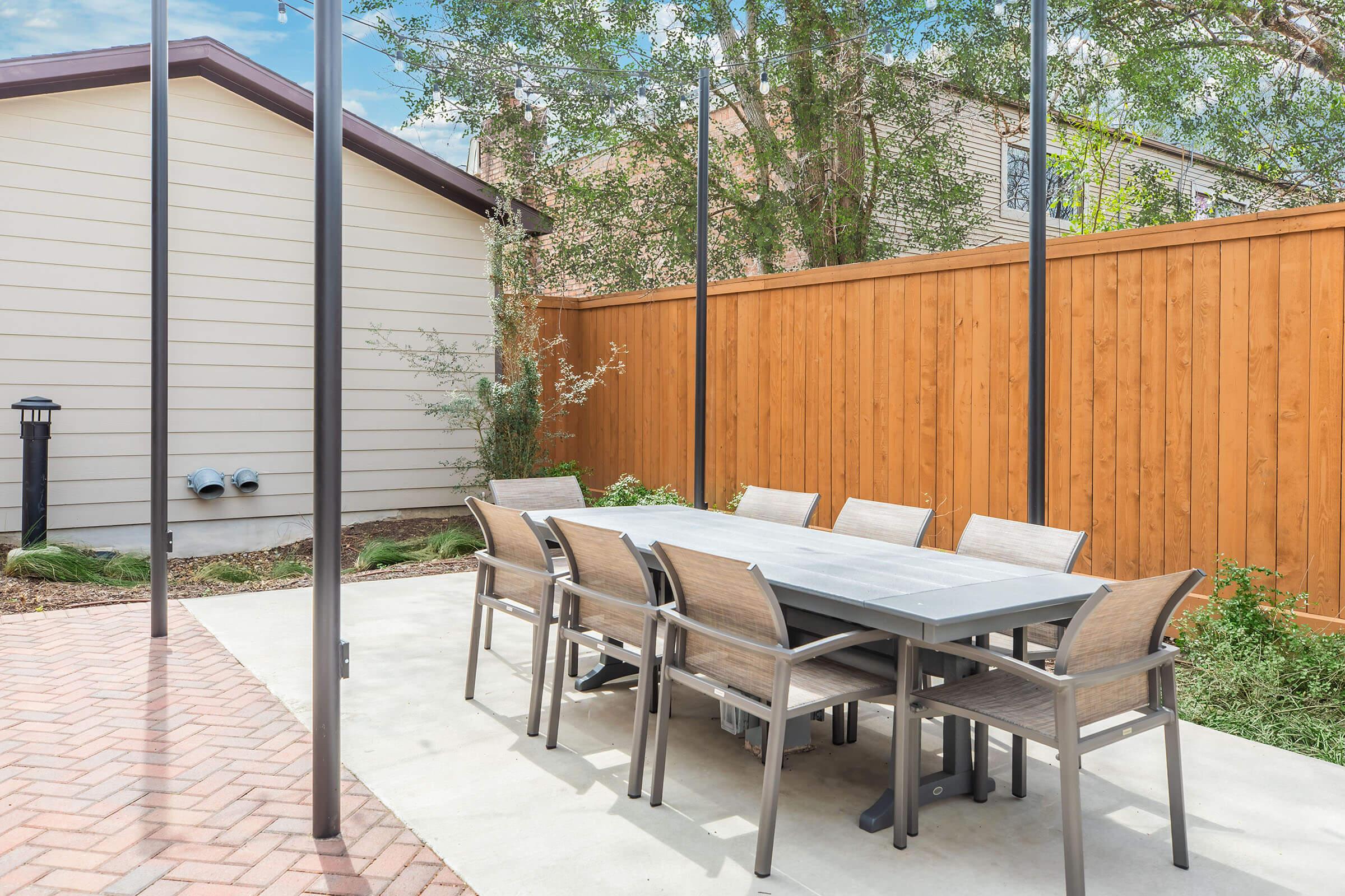 A spacious outdoor dining area featuring a long rectangular table with eight chairs, set on a paved patio. The surrounding area includes a wooden fence and some greenery, with a house visible in the background. The scene is bright and inviting, perfect for outdoor gatherings.