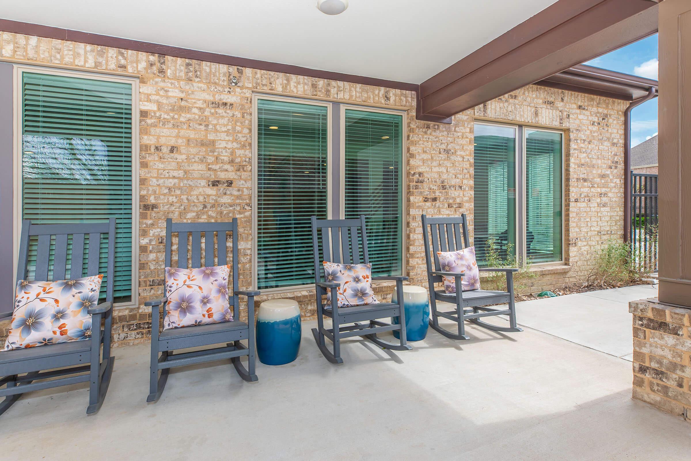 Outdoor seating area featuring four rocking chairs with floral cushions, positioned on a concrete patio. Large windows with green shutters adorn the brick wall behind. Two decorative side tables in varying colors complement the seating, creating a cozy and inviting atmosphere.