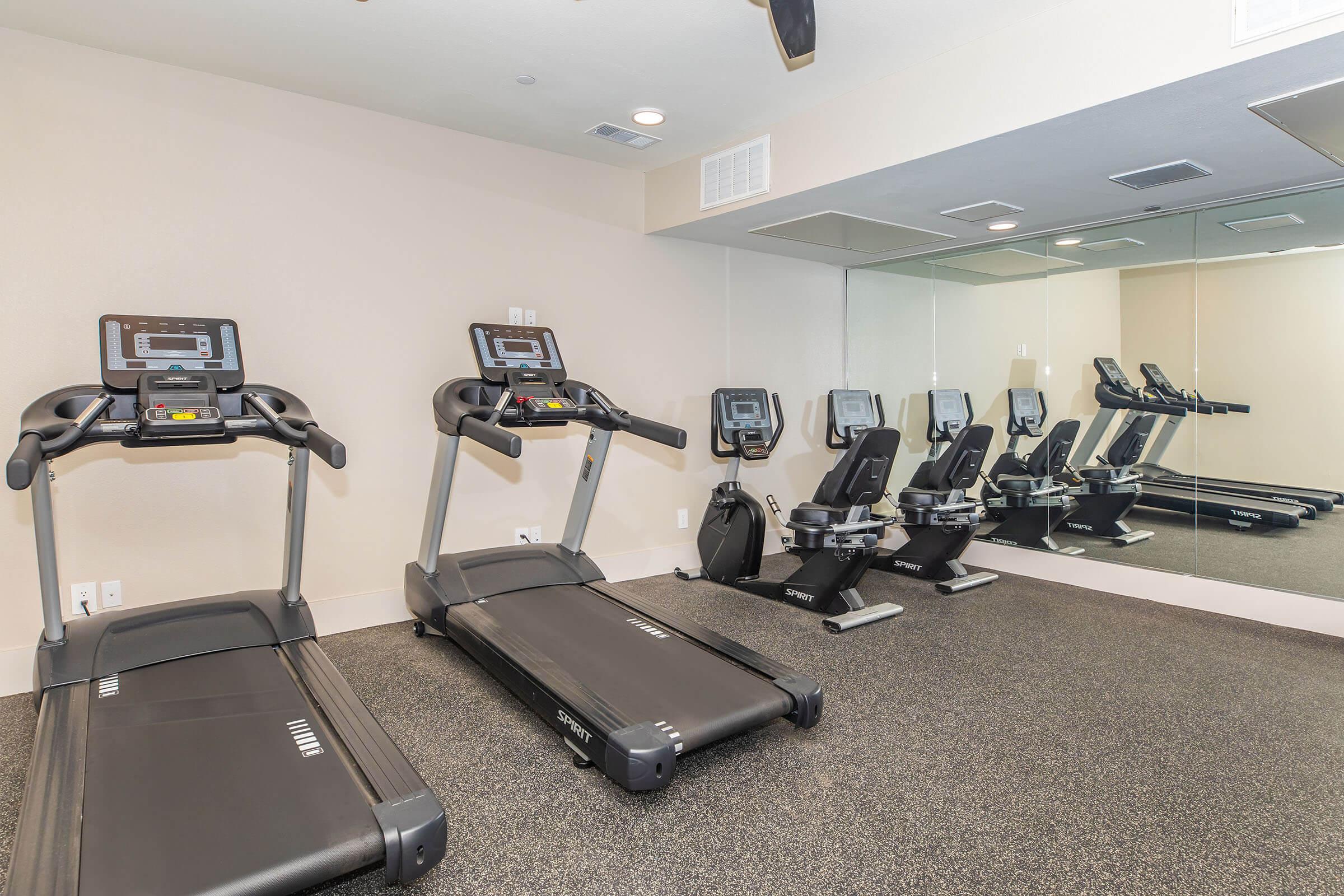 A clean, modern gym space featuring two treadmills and three stationary bikes. The flooring is black, and there is a large mirror on one wall, reflecting the equipment. The overall atmosphere is bright and inviting, ideal for a workout.