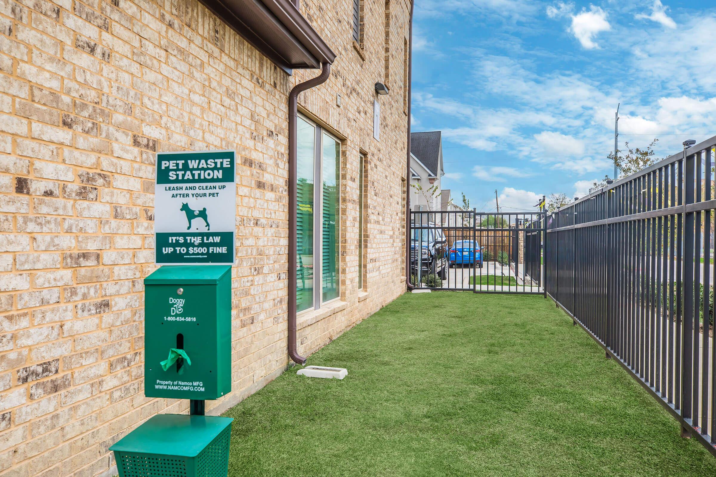 Pet waste station beside a fenced area, featuring a green disposal unit with instructions for pet owners to clean up after their dogs. The background shows a brick wall and a blue sky with some clouds.