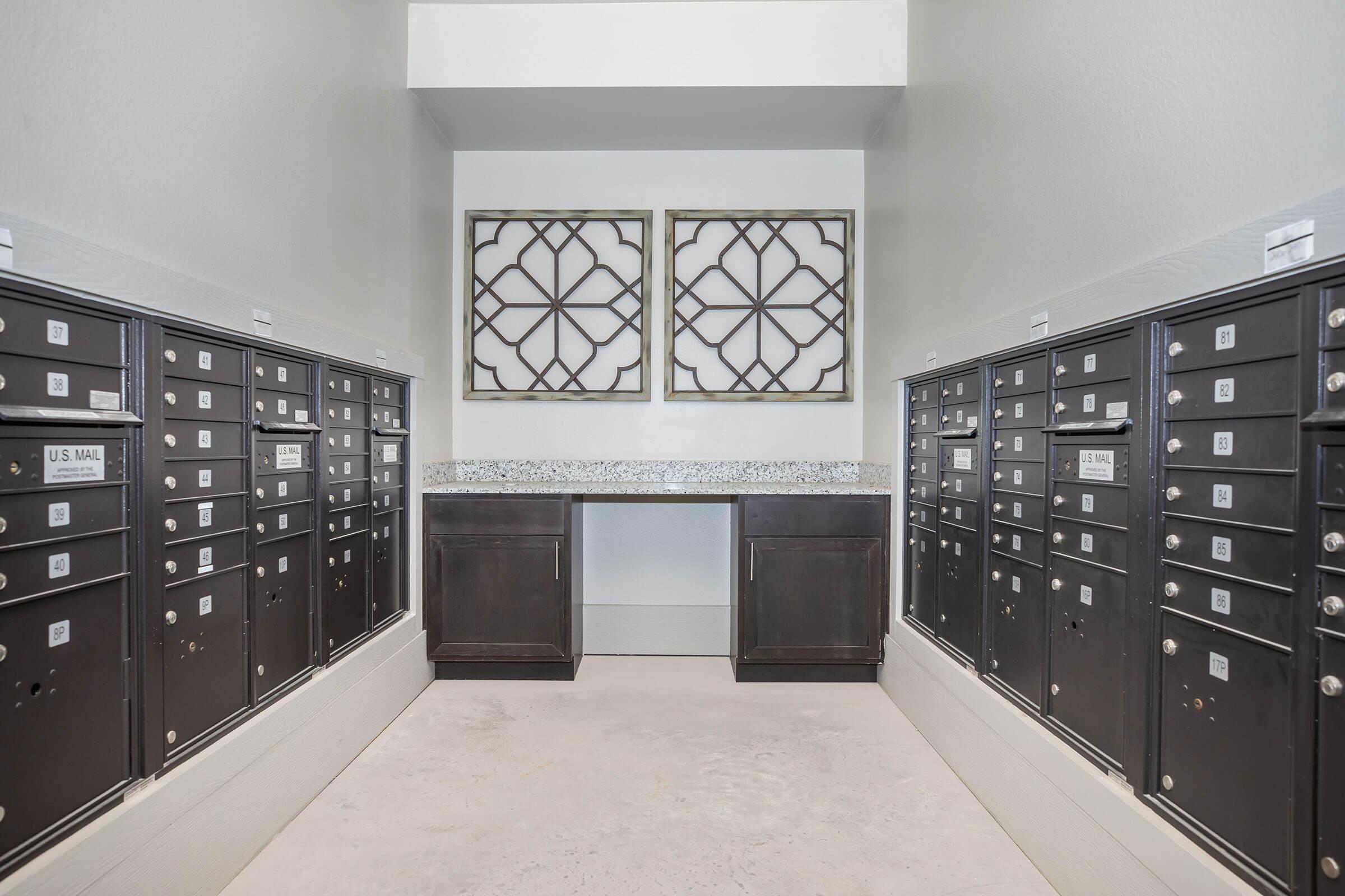 A modern interior featuring two rows of black mailboxes against a light wall. A dark wood counter with a granite top is centered between the rows, and two decorative framed wall art pieces are hung above. The floor is a neutral tone, creating a clean and organized mail area.