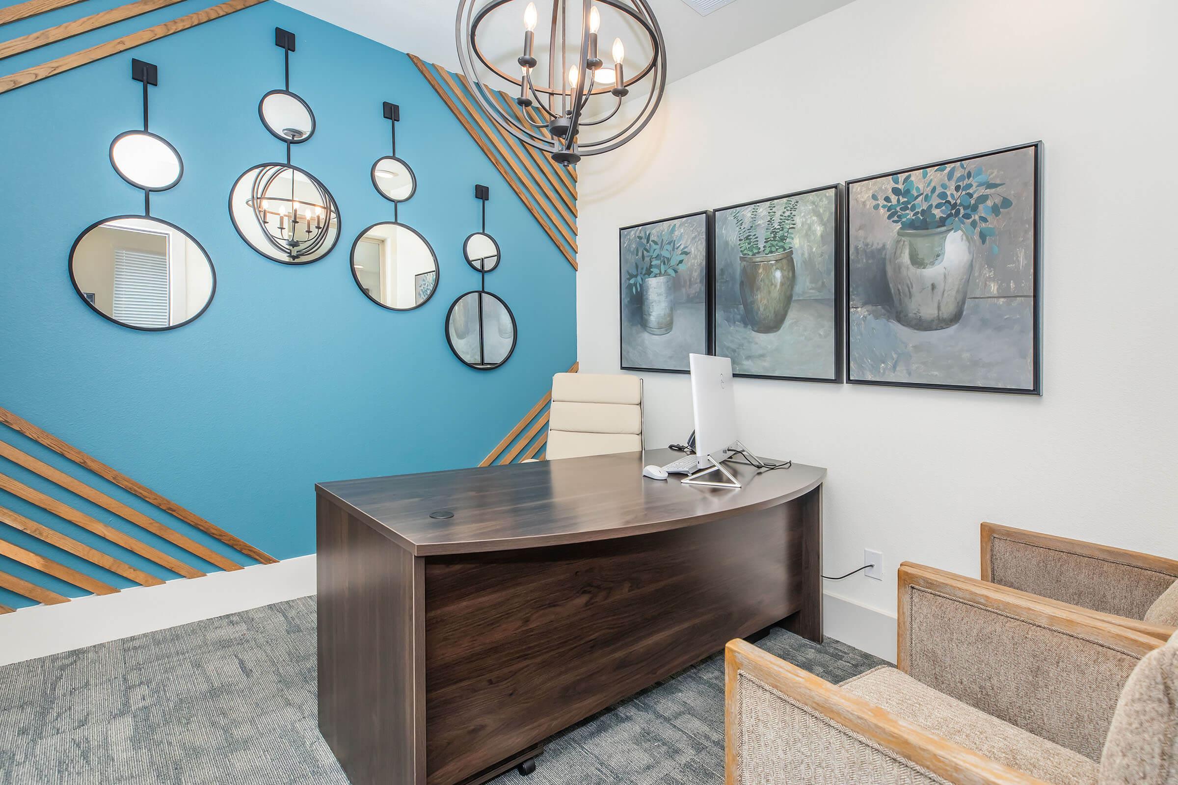 A modern office interior featuring a wooden desk with a computer, stylish beige chair, and two comfortable armchairs. The wall behind is painted blue with wooden accents and features multiple circular mirrors. Two framed artworks depicting potted plants are hung above the desk, enhancing the contemporary design.