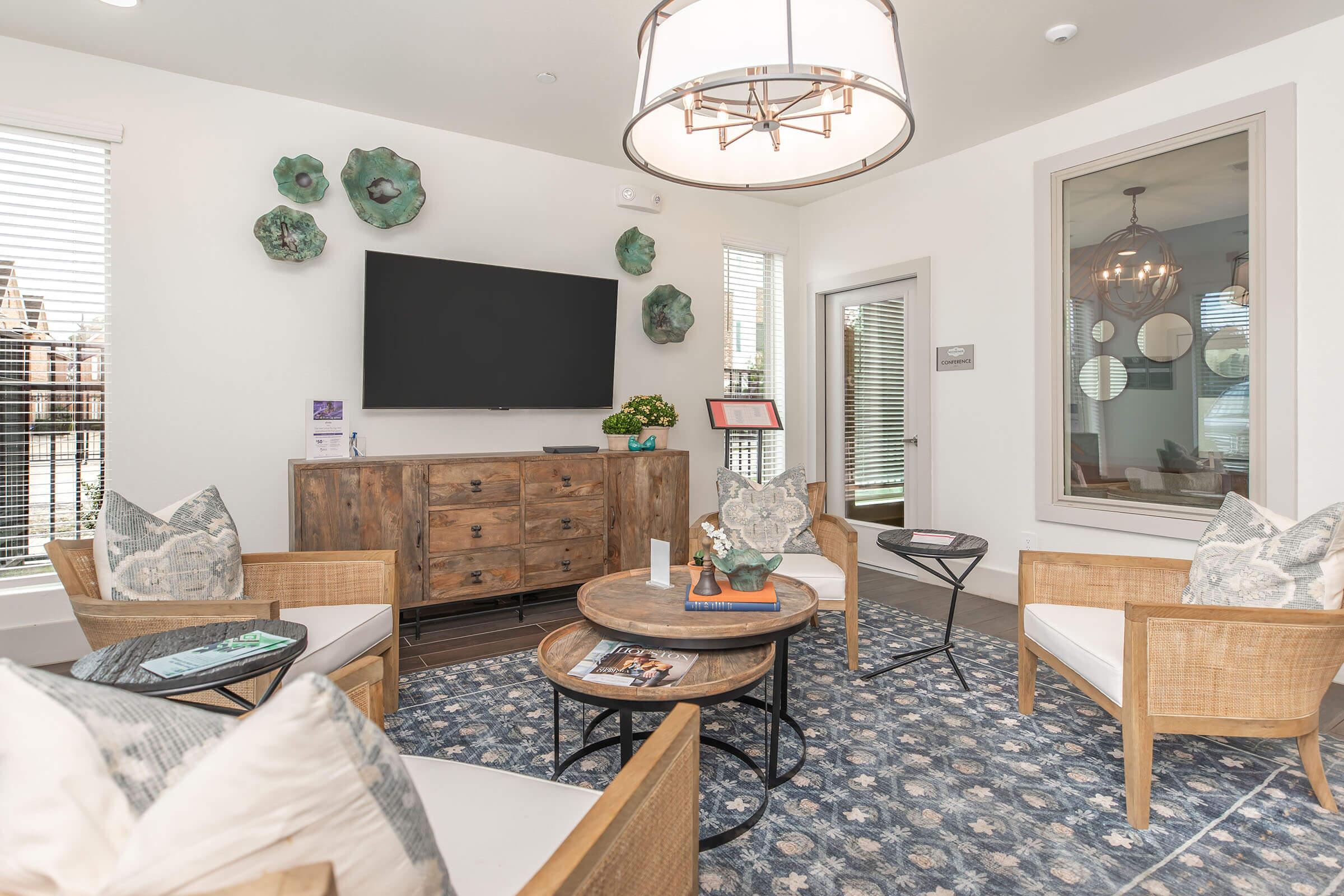 A modern living area featuring a wooden media console with a television, surrounded by stylish seating. Two wicker chairs with cushions sit opposite a round coffee table, while a patterned rug adds warmth. Decorative wall art and potted plants enhance the inviting ambiance of the space.