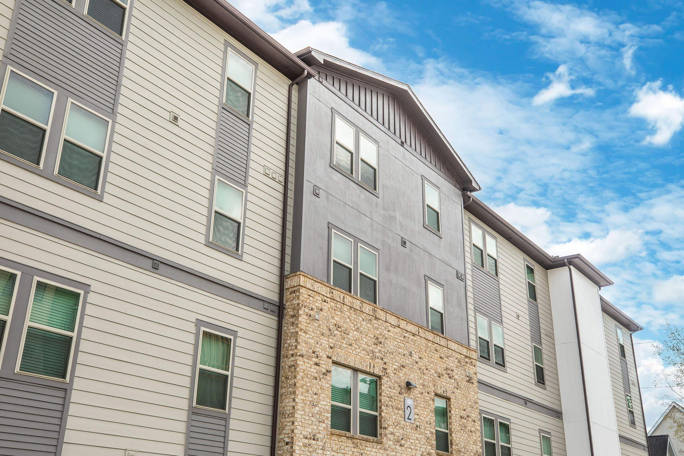 A multi-story apartment building with a mix of grey and stone exterior. The building features several windows and a modern architectural design, set against a bright blue sky with scattered clouds.