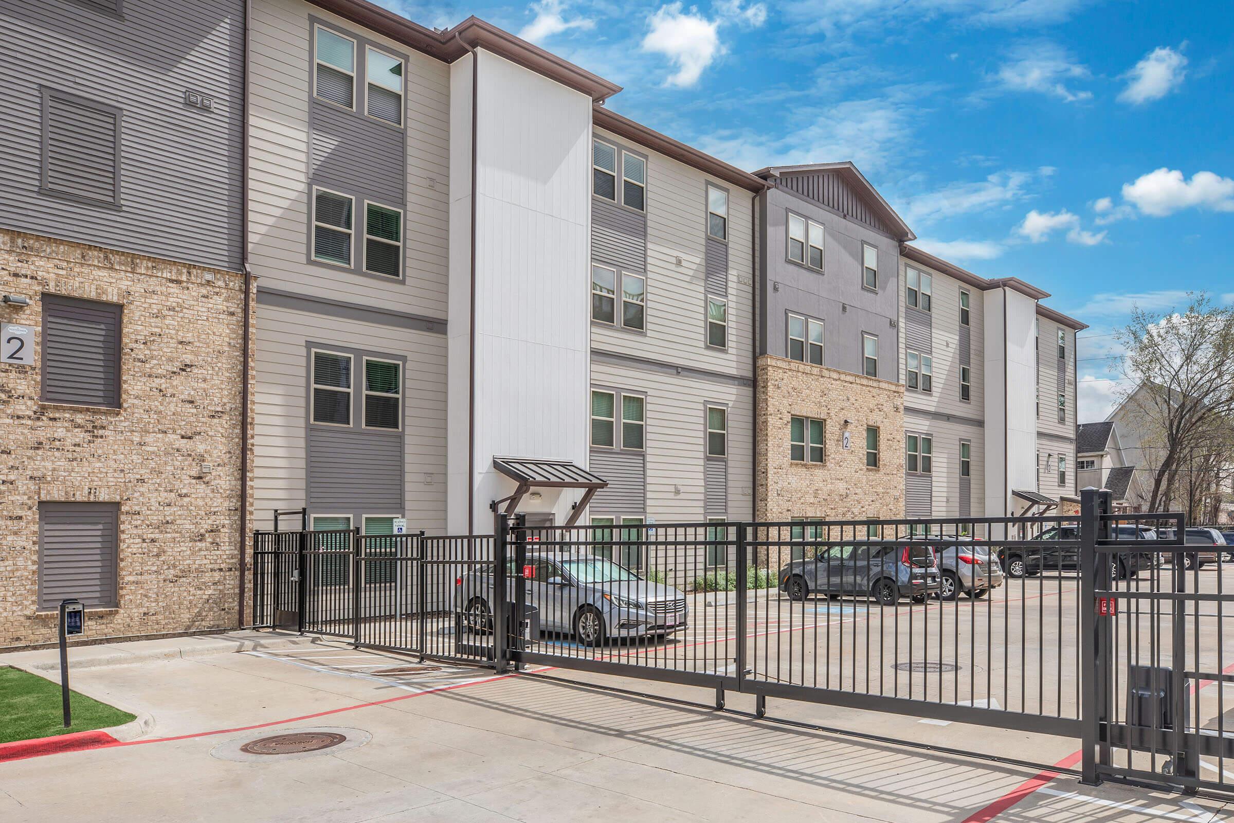 Residential apartment building with a mix of brick and siding exteriors, featuring multiple floors, large windows, and a gated entrance. Several parked cars are visible in the driveway. The sky is clear with a few clouds, indicating a bright day.