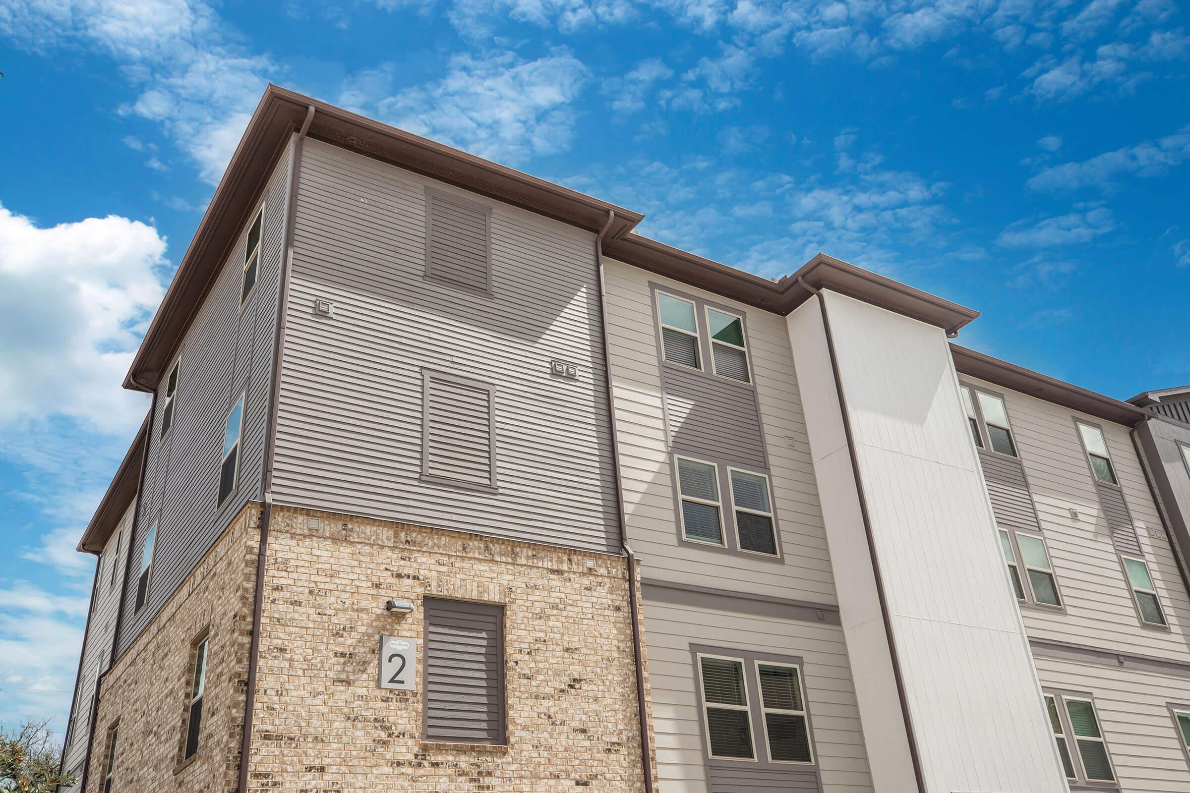 A modern multi-story apartment building featuring a mix of light gray siding and brick exterior. The building has multiple windows and a bold roofline, set against a bright blue sky with scattered clouds.