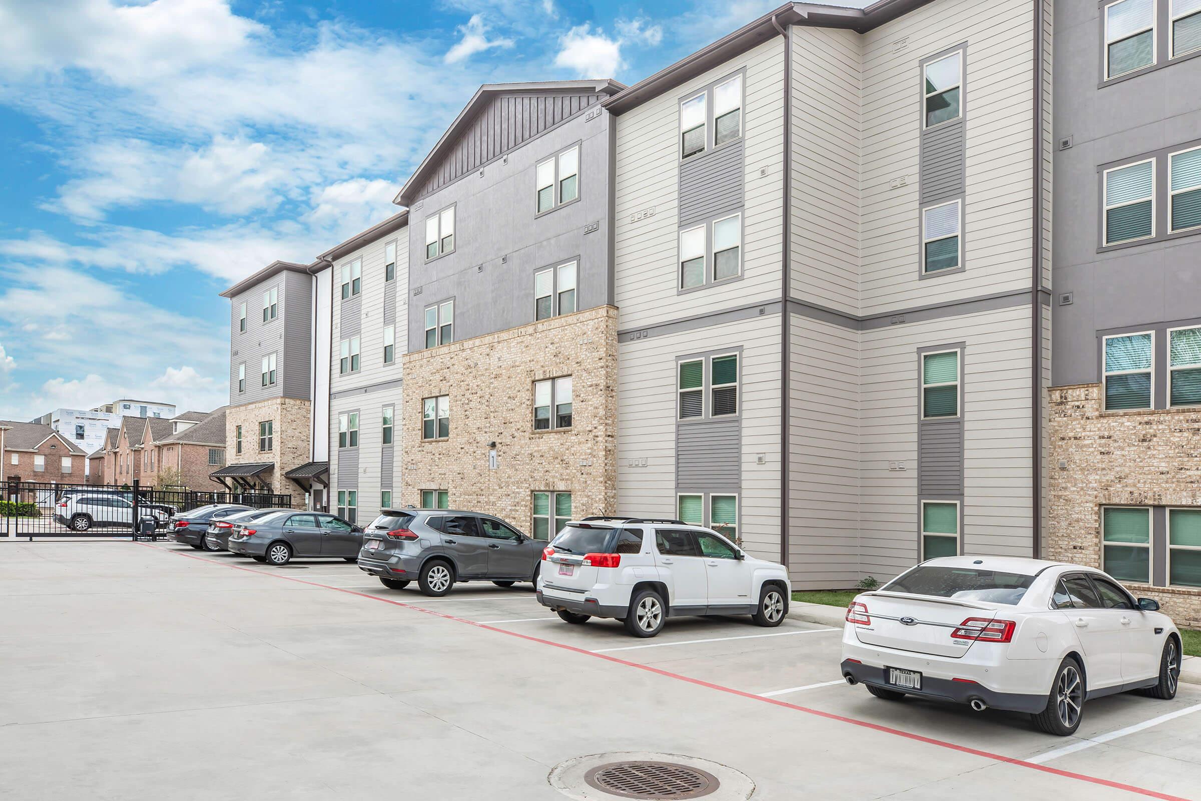 Residential apartment complex with a modern design featuring multiple floors, a mix of brick and siding exterior, and several parked cars in the foreground. The parking area is paved, and a blue sky with scattered clouds is visible above the building.