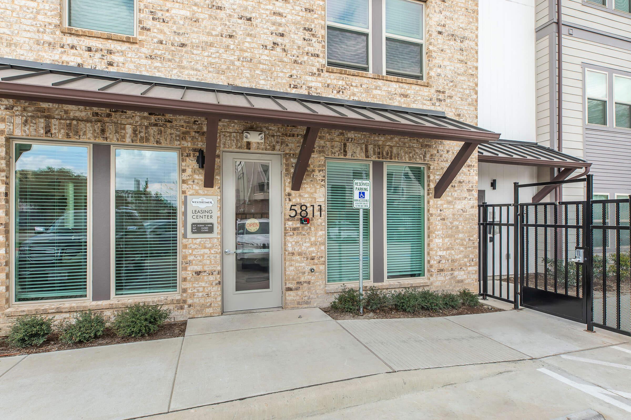 Exterior view of a building featuring a library center entrance with large windows, a welcoming door, and a sign displaying the address "5811." The facade is made of brick, and there are small plants and shrubbery along the pathway. A gated area is visible beside the entrance.