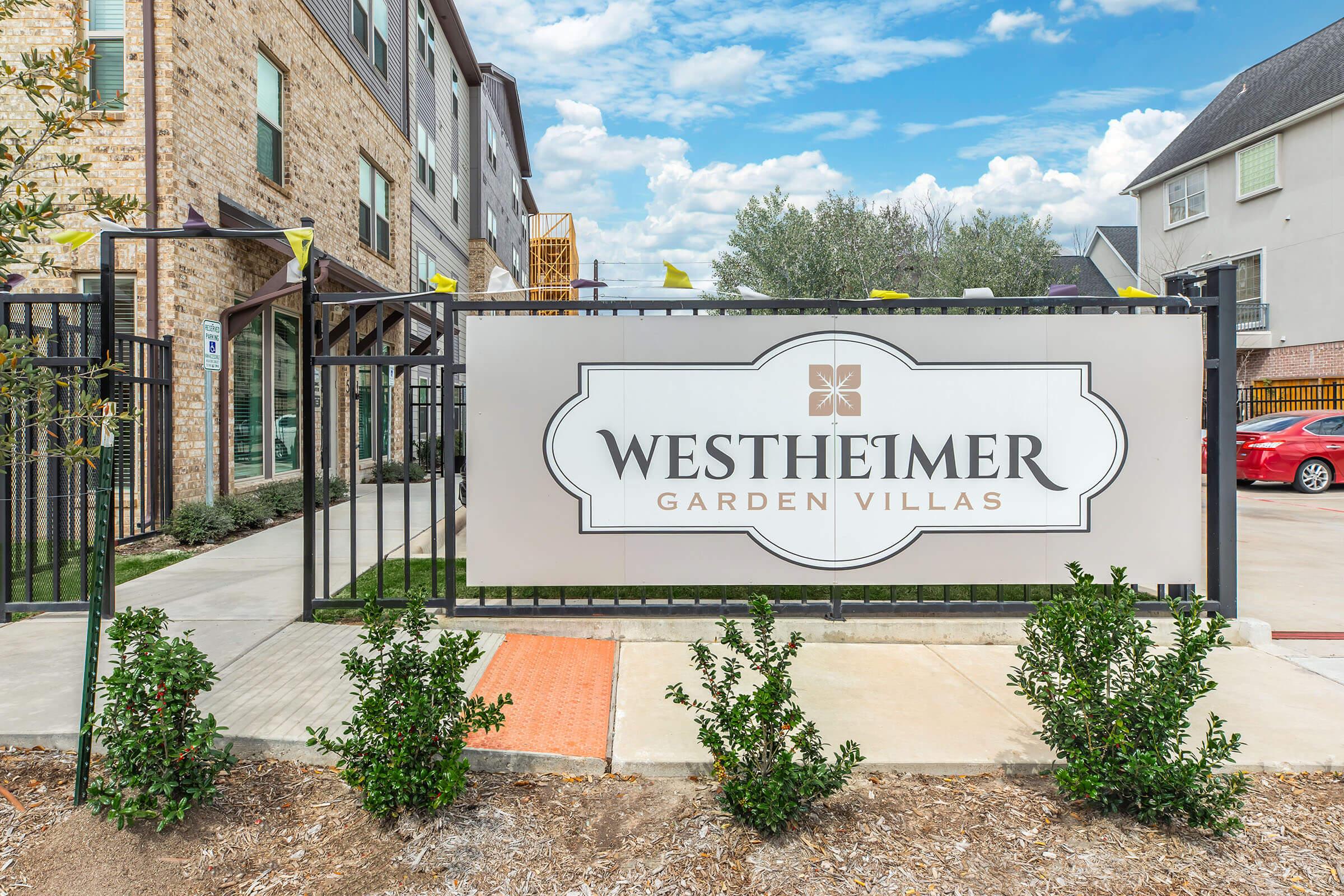 Sign for Westheimer Garden Villas, featuring the name prominently in a decorative font. The sign is surrounded by manicured shrubs and is located at the entrance of a modern residential building complex. The background shows a clear sky and contemporary architecture.