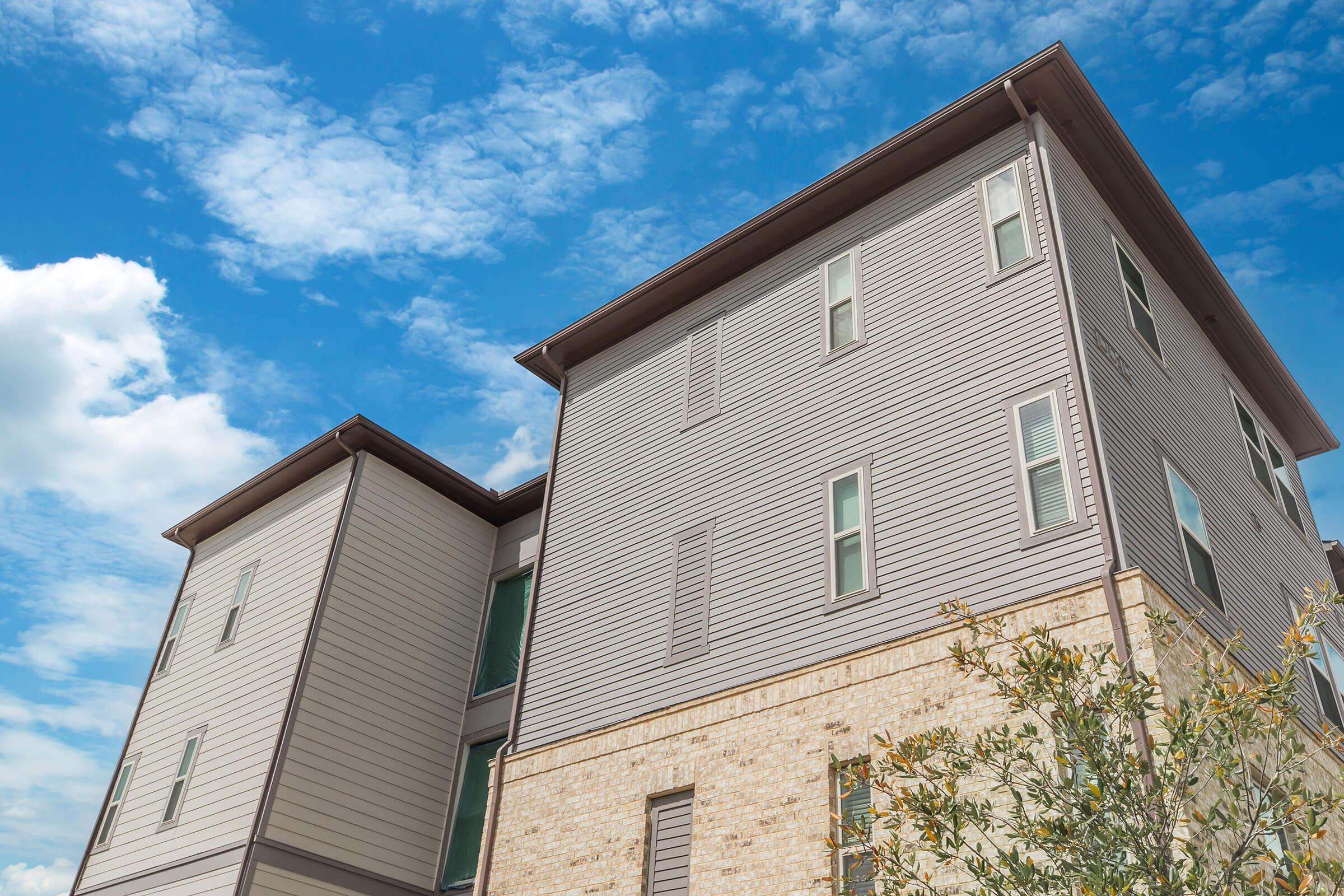 A modern two-story building with a combination of gray siding and light-colored brick. The structure has multiple windows and is set against a backdrop of a partly cloudy blue sky. There is a small tree or shrub in the foreground, adding a touch of greenery to the scene.