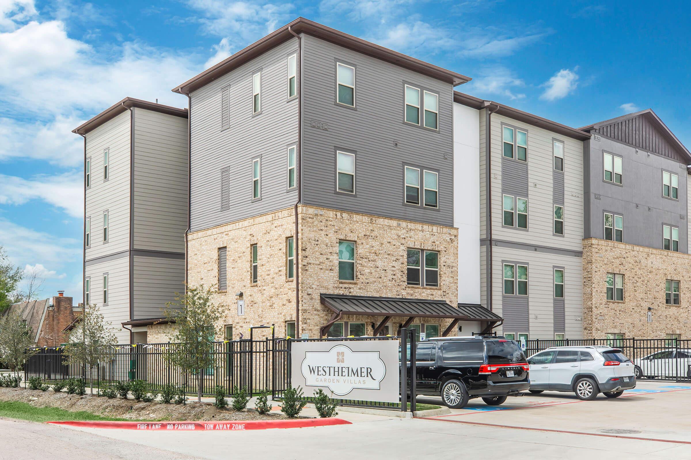 Modern multi-story apartment building featuring a mix of gray siding and stone accents, surrounded by a landscaped area and a gated parking lot with vehicles. The entrance is marked with a sign that reads "Westheimer," indicating the name of the property. Clear blue sky adds to the welcoming atmosphere.