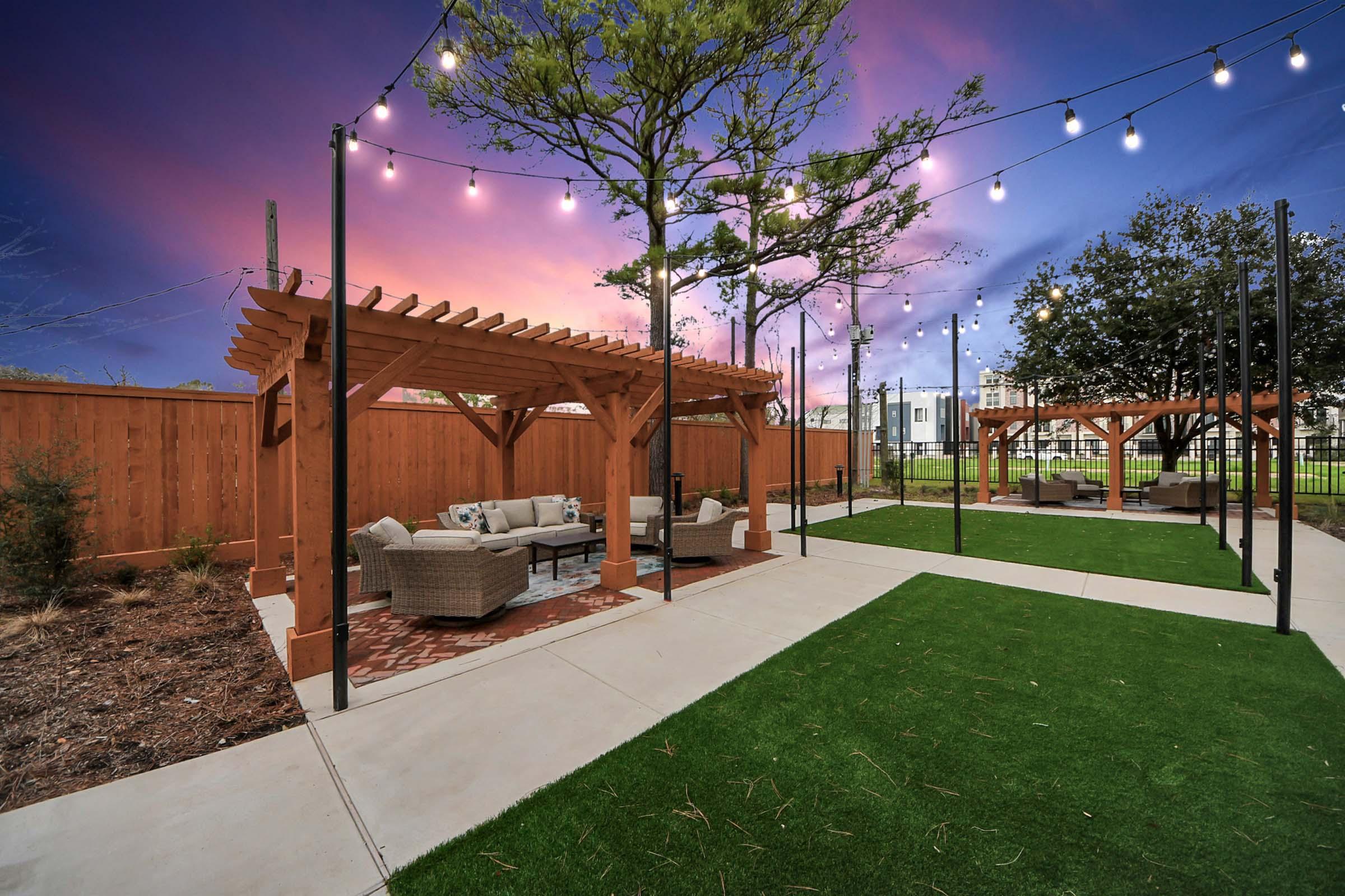 A serene outdoor space featuring two wooden pergolas with cozy seating arrangements. The area is surrounded by lush green grass and illuminated by string lights against a vibrant sunset sky, creating a warm and inviting atmosphere for relaxation and gatherings.