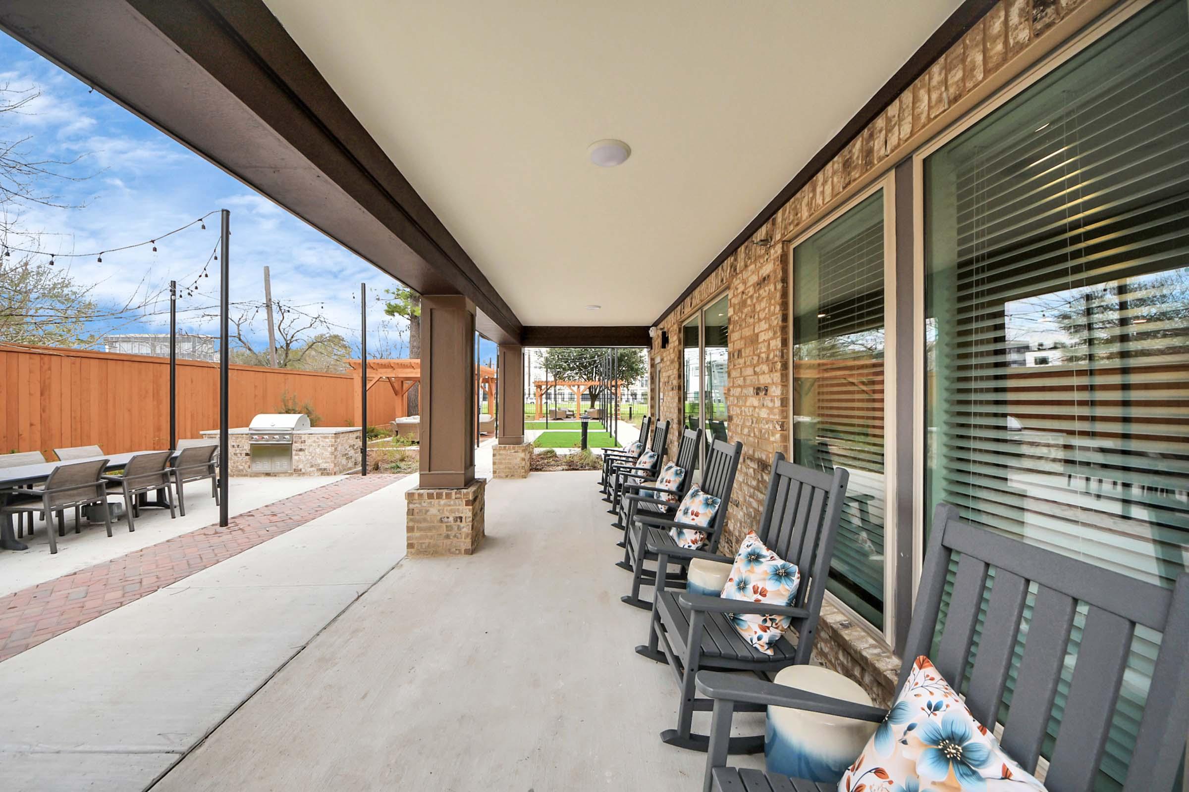A covered porch area featuring several rocking chairs with decorative pillows. In the background, there is a well-maintained lawn and patio set, surrounded by a wooden fence and landscaping. The sky is partly cloudy, creating a relaxed outdoor atmosphere.