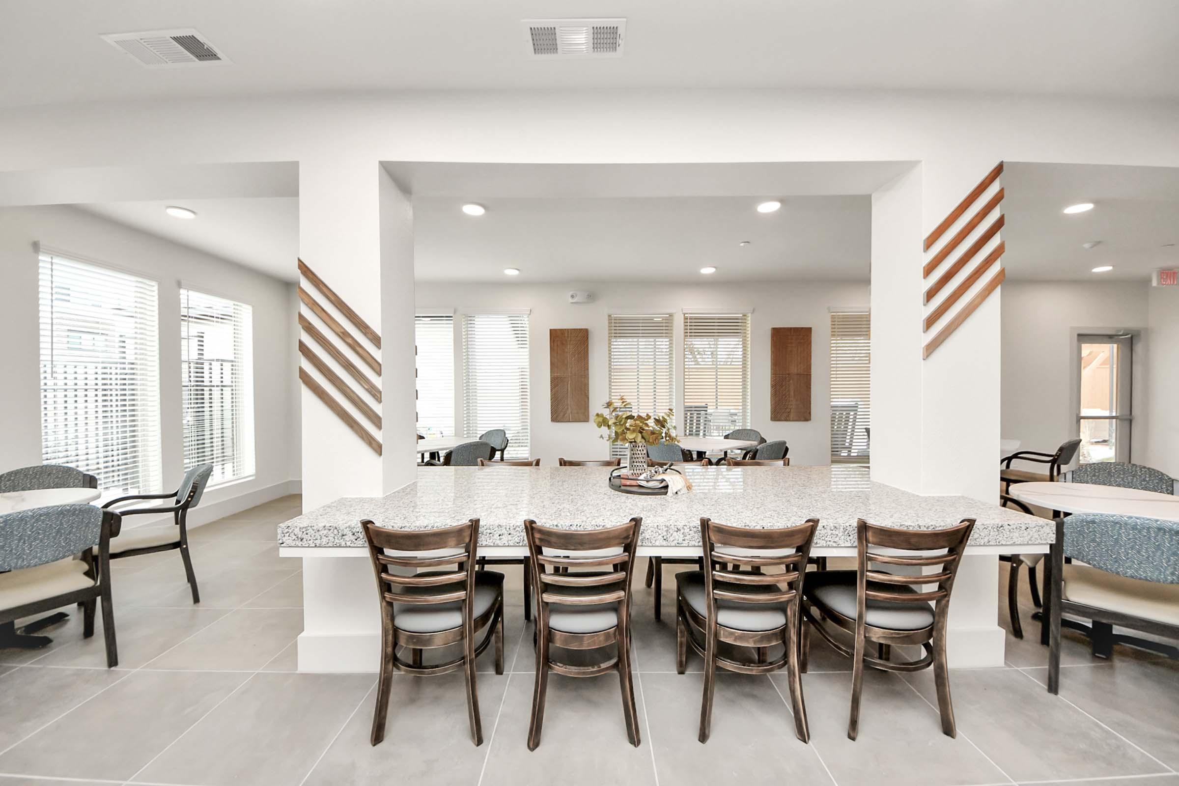A modern communal space featuring a long granite counter with wooden bar stools, surrounded by large windows allowing natural light. The room has a contemporary design with decorative wooden accents and seating areas, creating an inviting atmosphere for residents or guests.