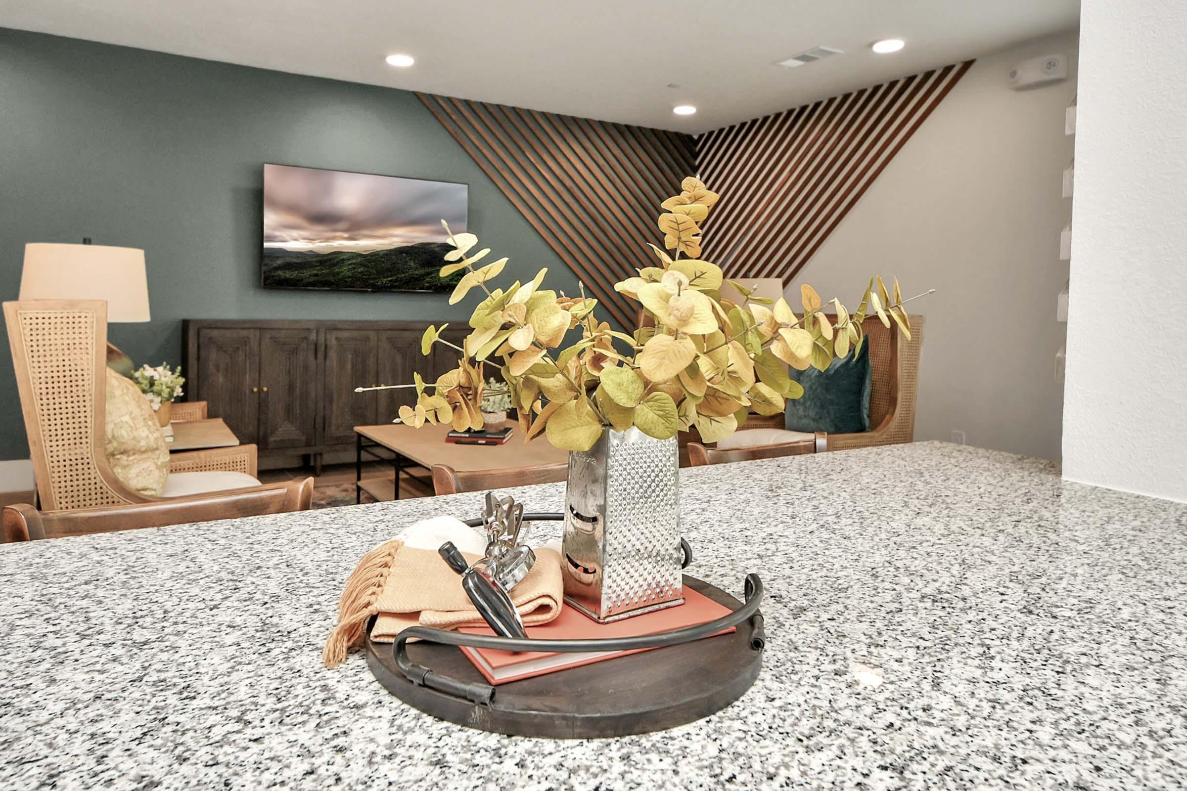 A stylish living space featuring a central decorative arrangement of dried flowers in a metallic vase on a countertop. The background includes a modern TV mounted on a wall, a wooden sideboard, and comfortable seating. The decor has a contemporary design with textured elements and earthy colors.