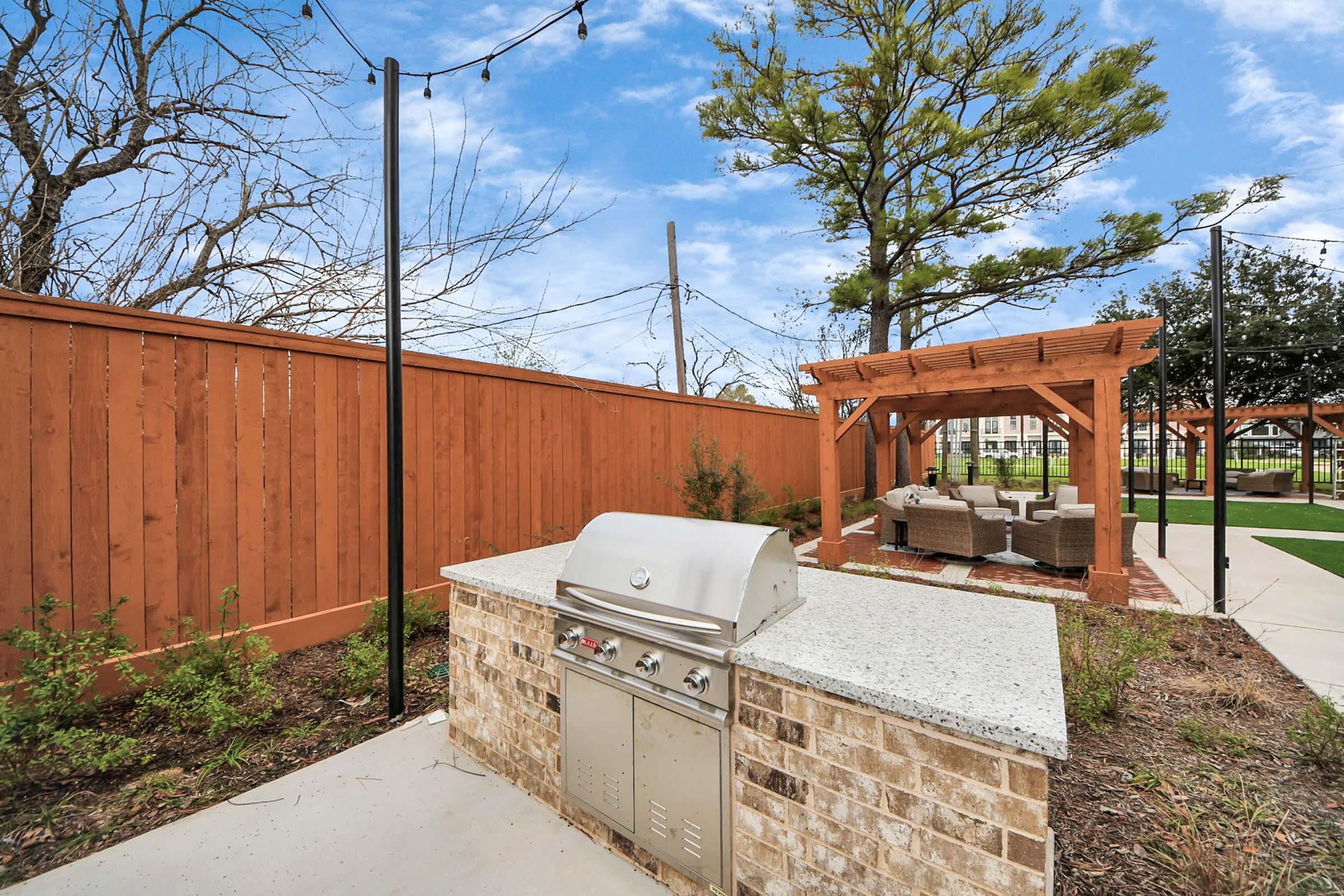 An outdoor kitchenette featuring a built-in grill and stone countertop, surrounded by a well-maintained patio area. In the background, there are wooden fences and a decorative pergola with seating, complemented by green grass and a clear blue sky.