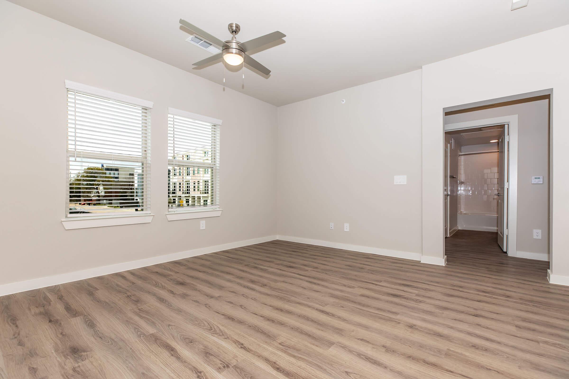 Interior of a modern, empty room featuring light-colored walls, large windows with blinds, a ceiling fan, and laminate flooring. A doorway on the right leads to another space, likely a bathroom or hallway. The overall ambiance is bright and spacious.