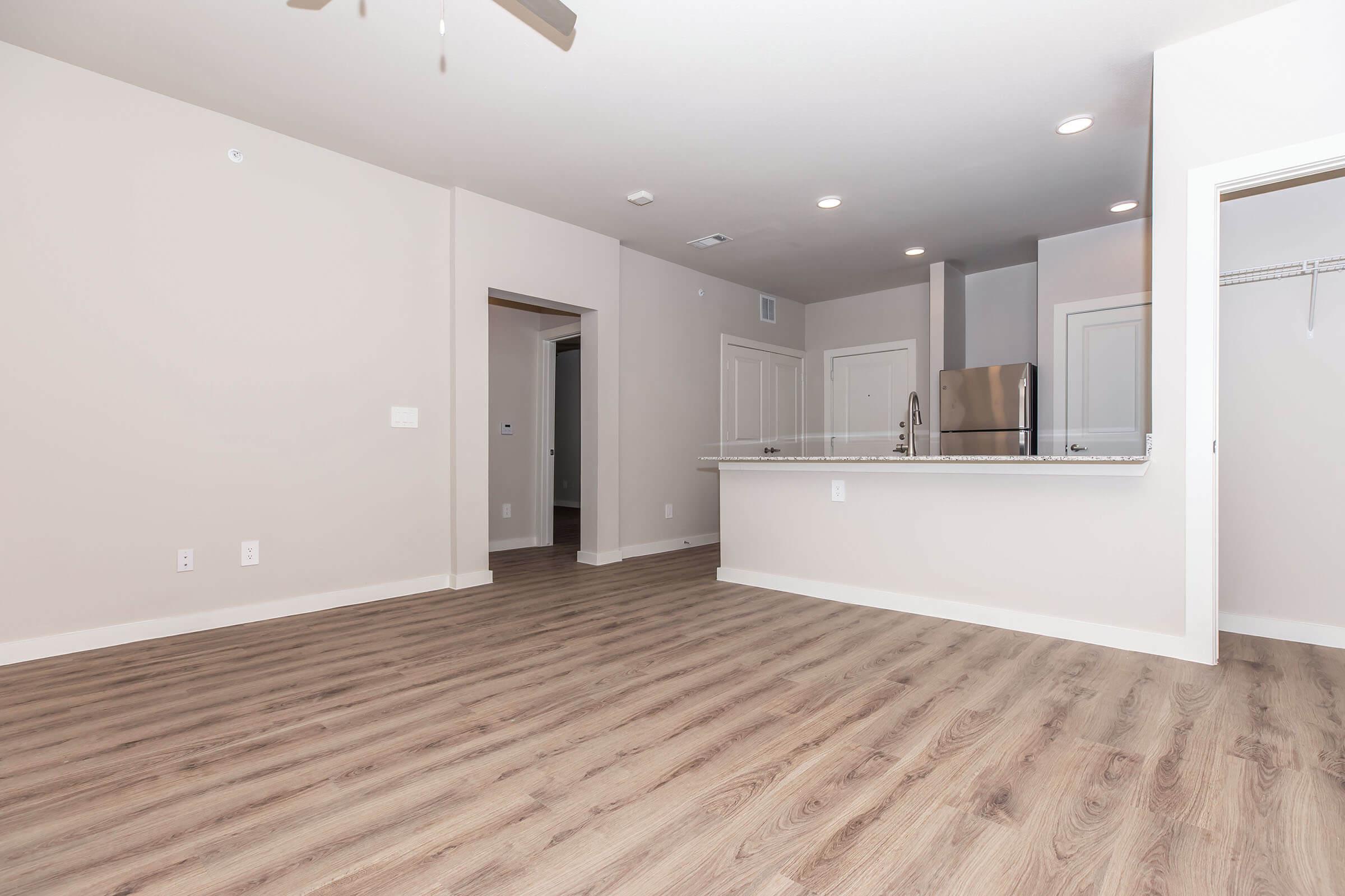 A modern, unfurnished living space featuring light-colored walls and wood-like flooring. The room includes a kitchen area with stainless steel appliances and a granite countertop, a ceiling fan, and doorways leading to other rooms. Natural light illuminates the space from multiple sources.
