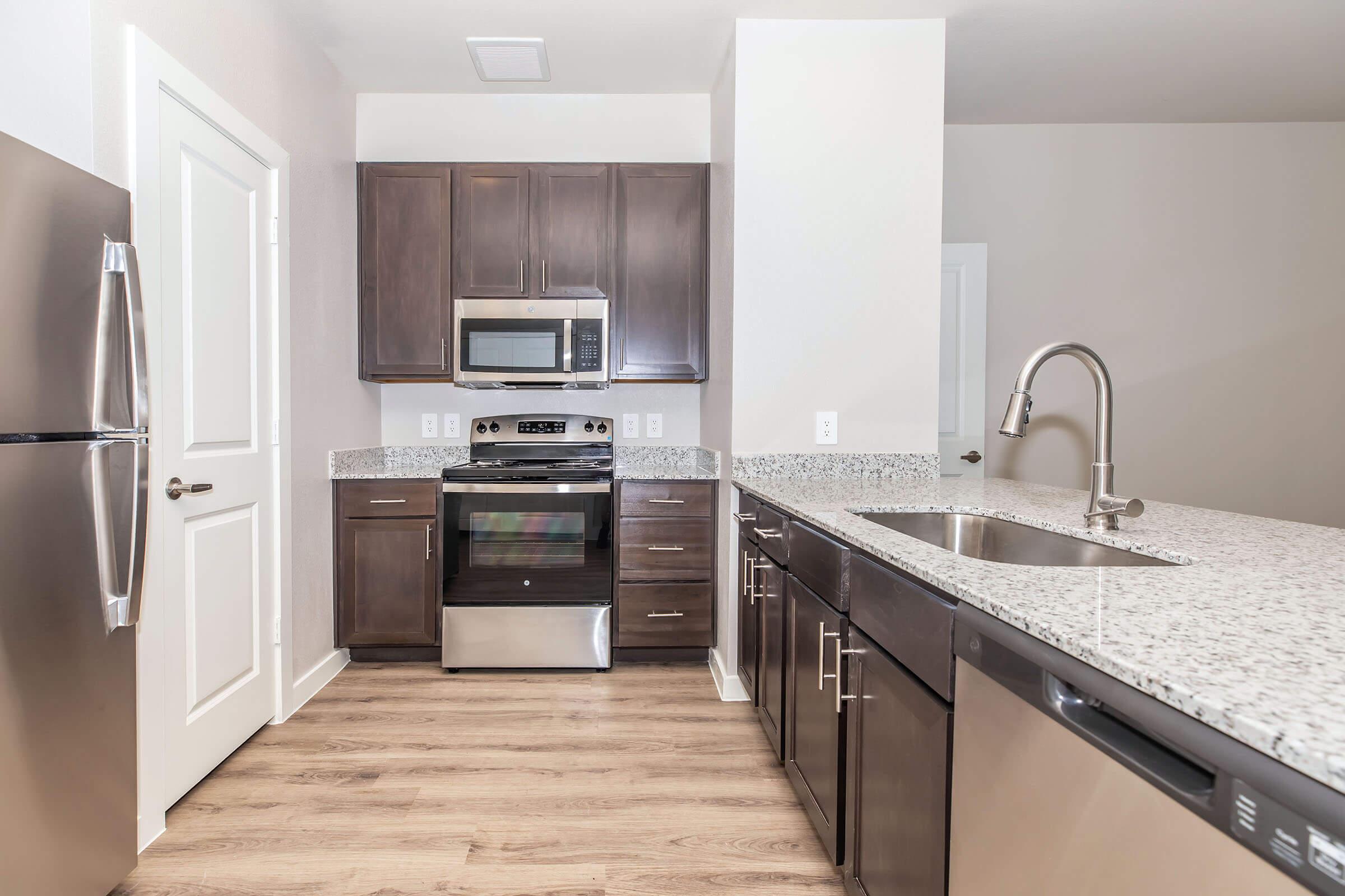 Modern kitchen featuring dark wood cabinets, stainless steel appliances, and a granite countertop. The space includes a built-in oven, microwave, and a sink with a sleek faucet. The flooring is a light wood laminate, and the walls are painted in neutral tones, creating a bright and inviting atmosphere.