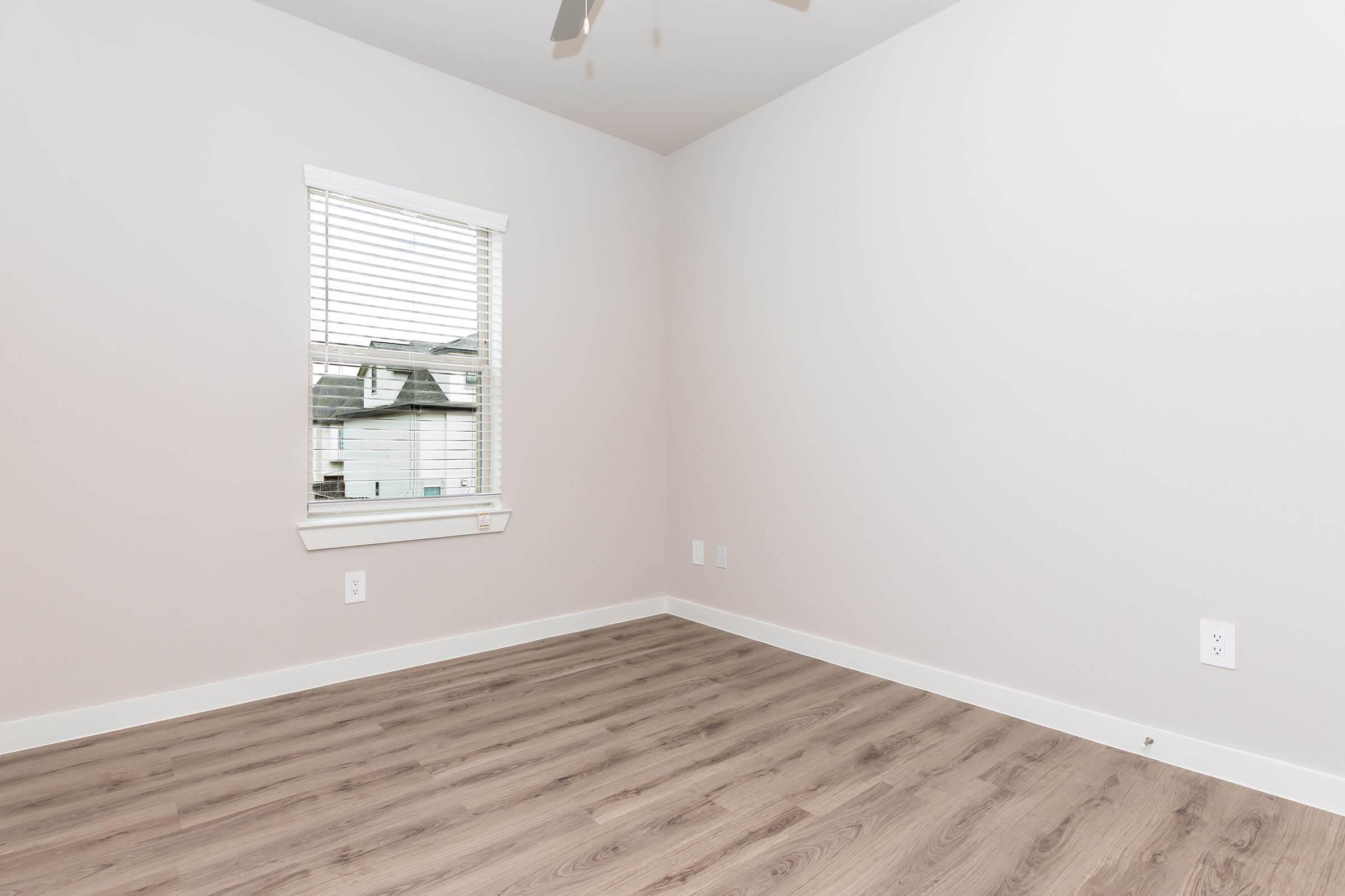 Empty room with light-colored walls and a wood-like floor. There is a window with blinds allowing natural light, framed by a thin white wall. A ceiling fan is visible above, and there are electrical outlets on the wall. The space is minimalistic and unfurnished.