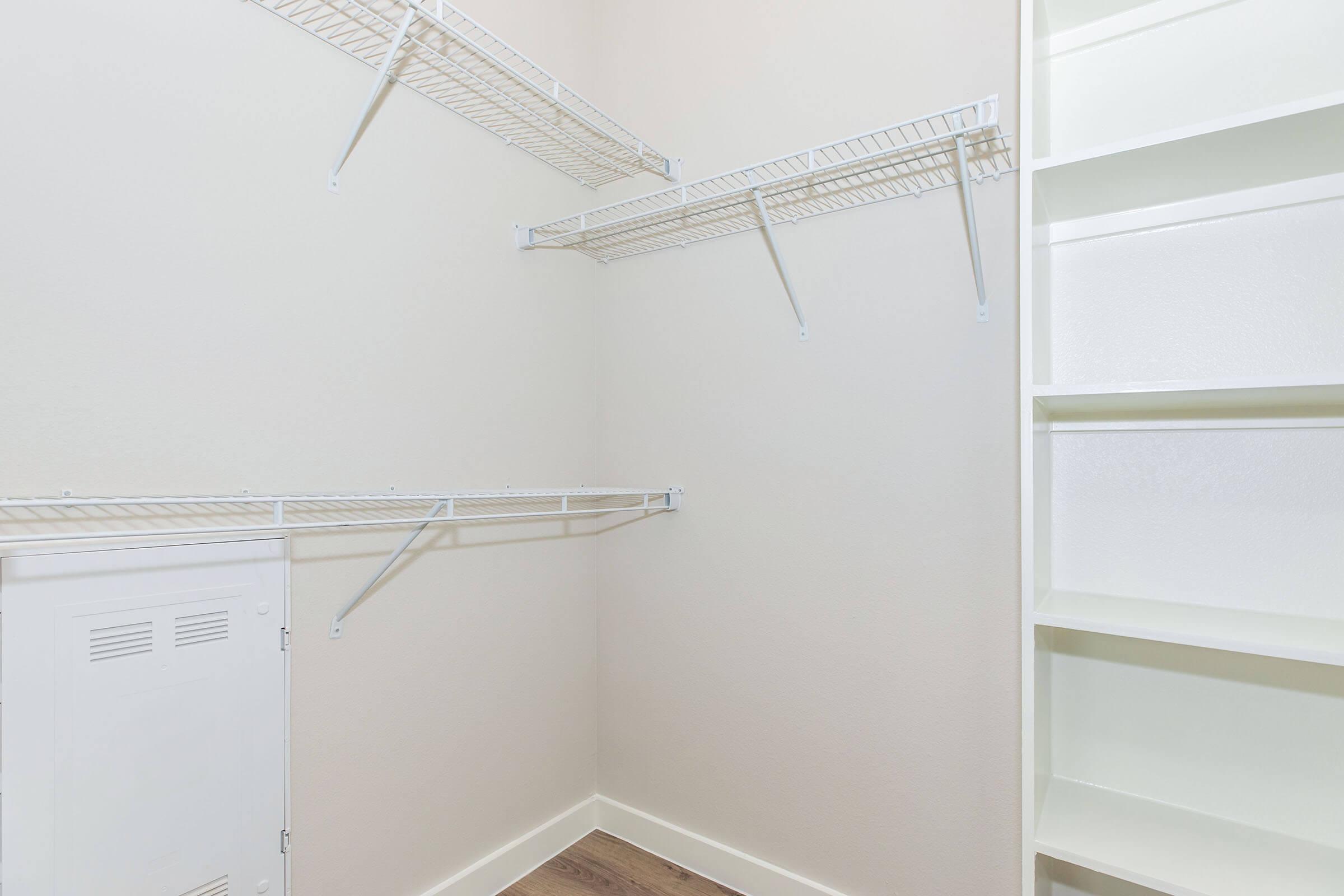 Empty closet with white wire shelving on two walls and a small utility door. The walls are painted a light color, and there's a built-in shelving unit on the right. The floor features wood-like flooring, contributing to a clean and organized appearance.