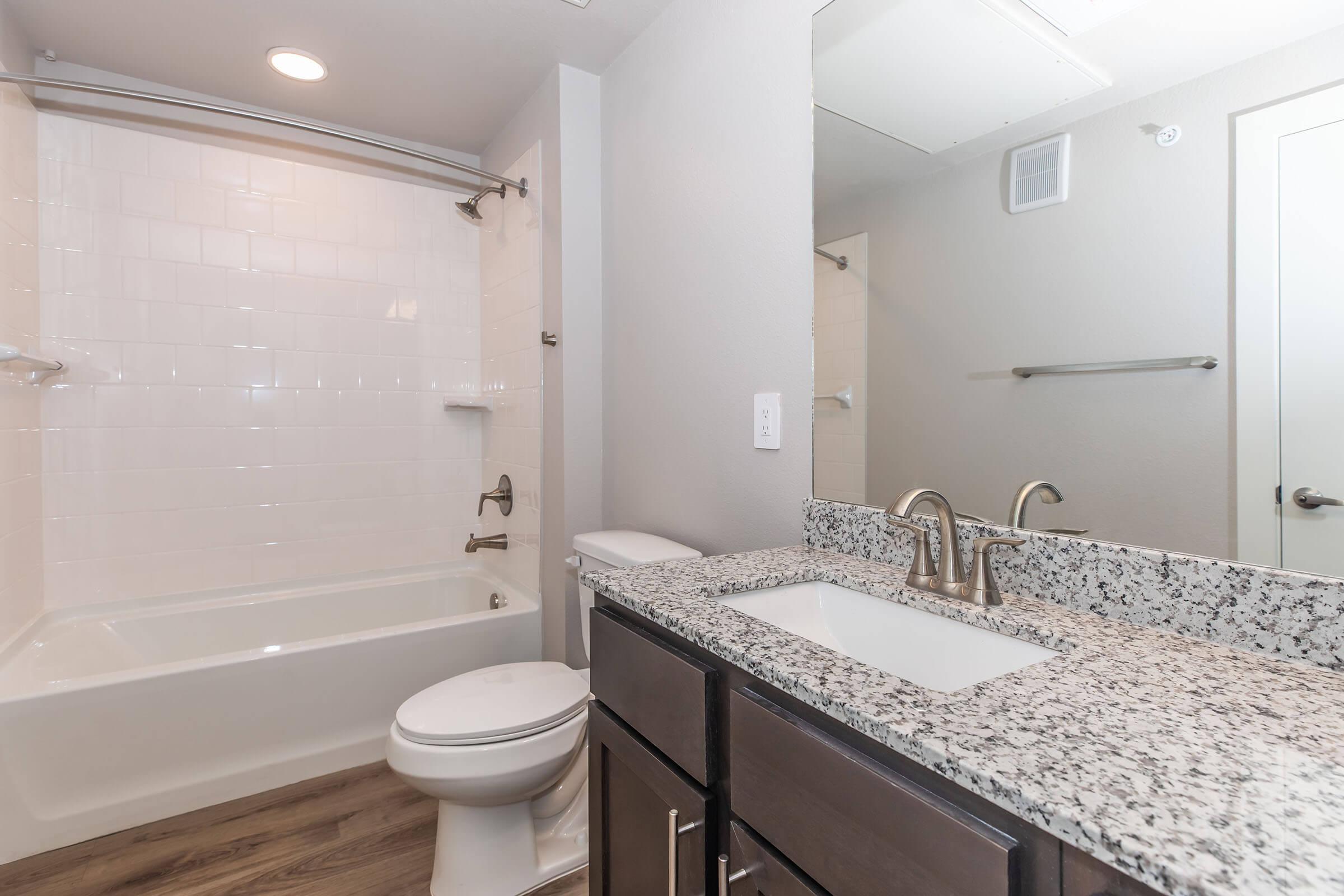 A modern bathroom featuring a white tiled shower and bathtub, granite countertops, a sink with contemporary faucets, and dark wood cabinetry. The space is well-lit with overhead lighting and includes a mirror on the wall.