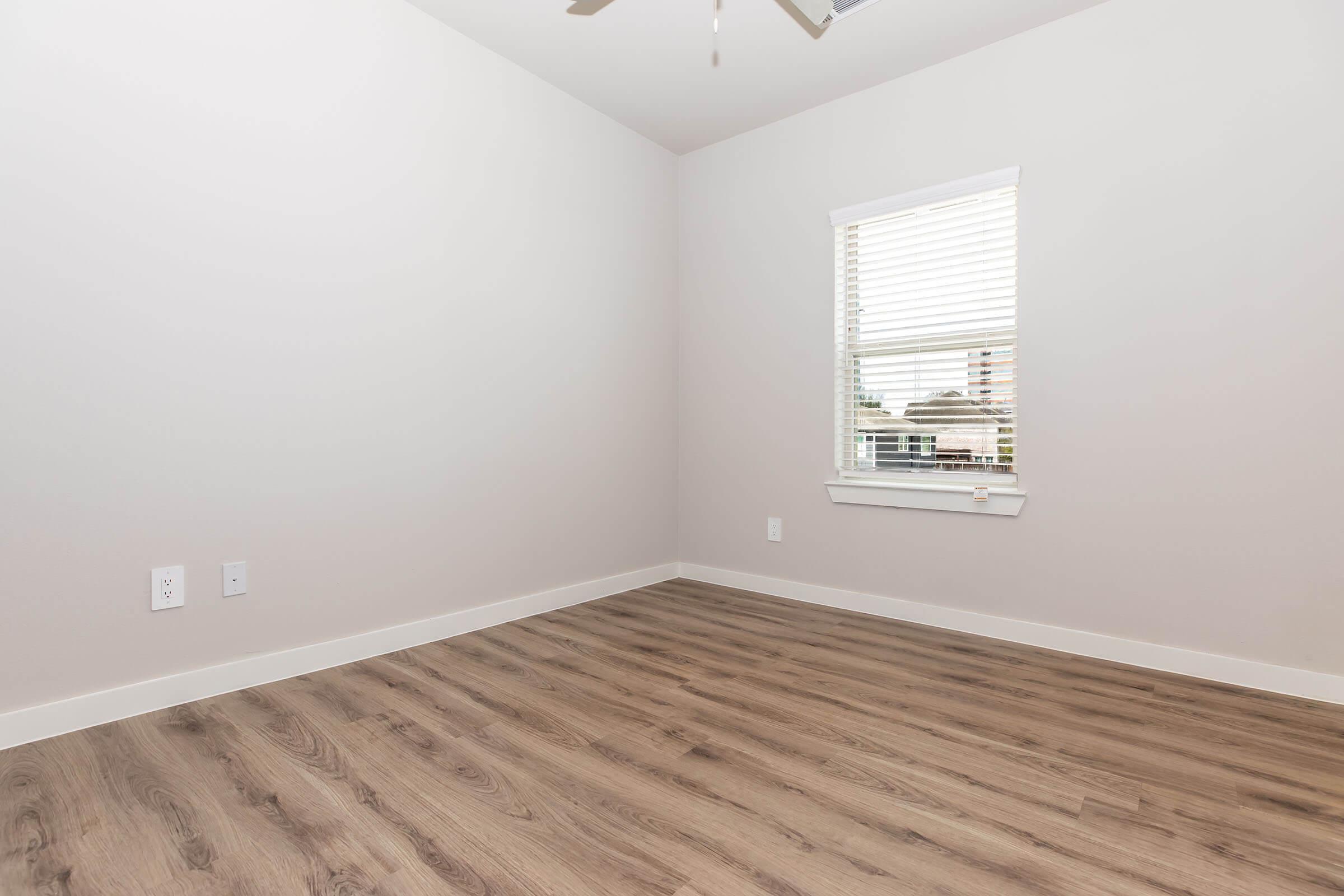 A spacious, empty room with light gray walls and a wood-like floor. A window with white blinds allows natural light to enter, and the ceiling features a ceiling fan. There are two electrical outlets on one wall, creating a minimalist and clean aesthetic.