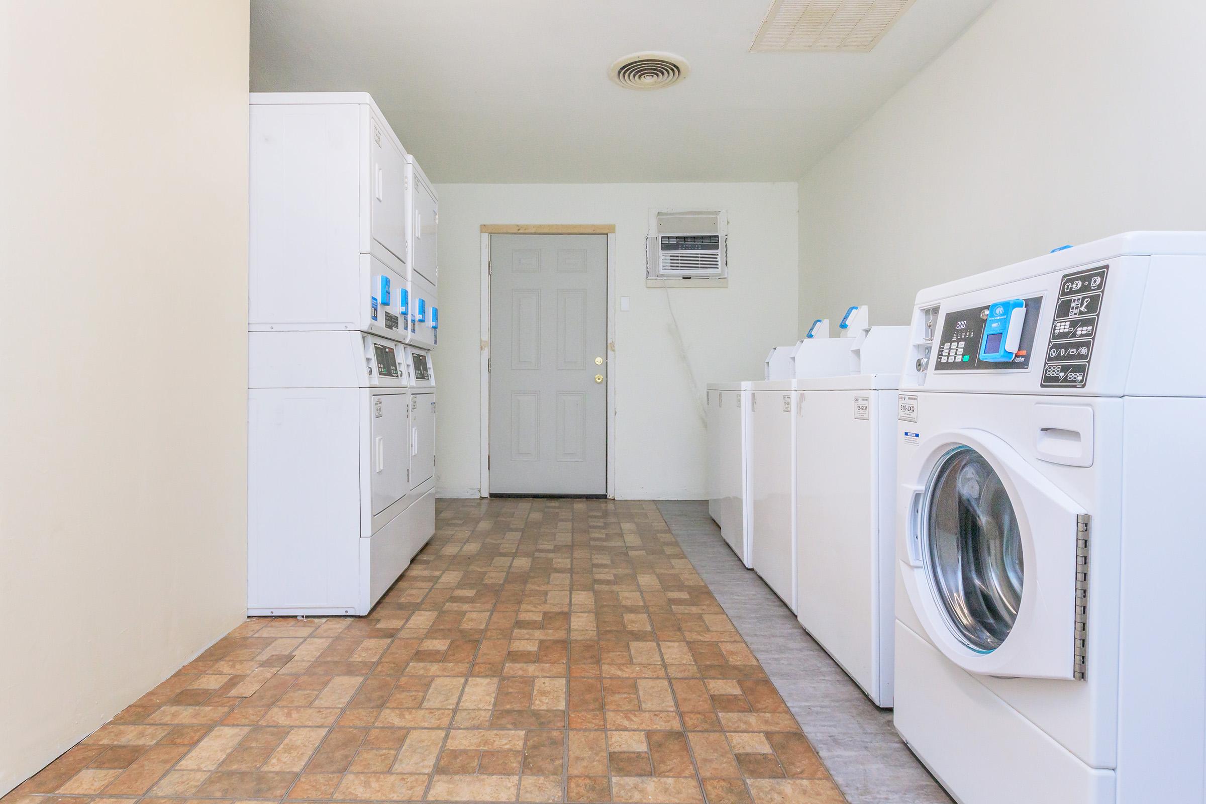 A clean and bright laundry room featuring several white washing machines and dryers lined against the walls, with a door at the far end. The floor has a patterned tile design, and there is an air conditioning unit visible above the door. The space is well-lit and organized.