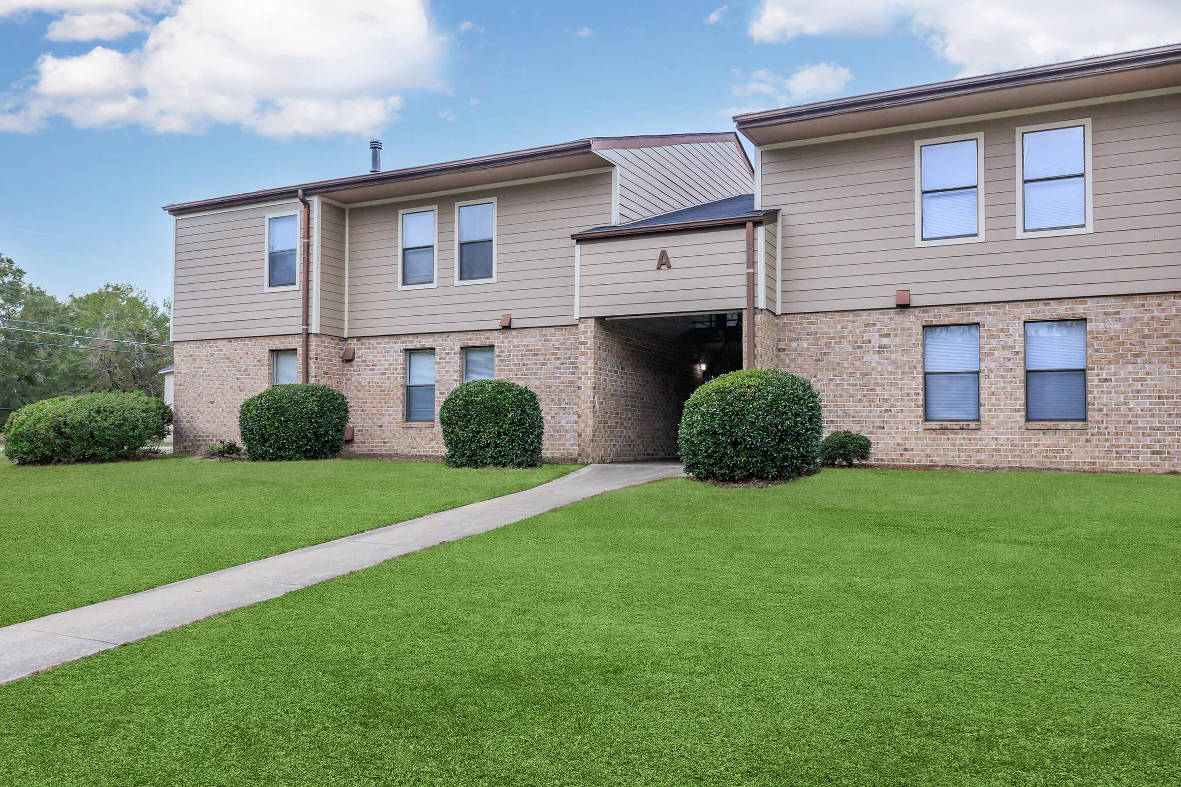A brick apartment building with green trimmed hedges and a well-maintained lawn. The entrance features a lightly colored overhang and a walkway leading into the building. The sky is clear with a few clouds.