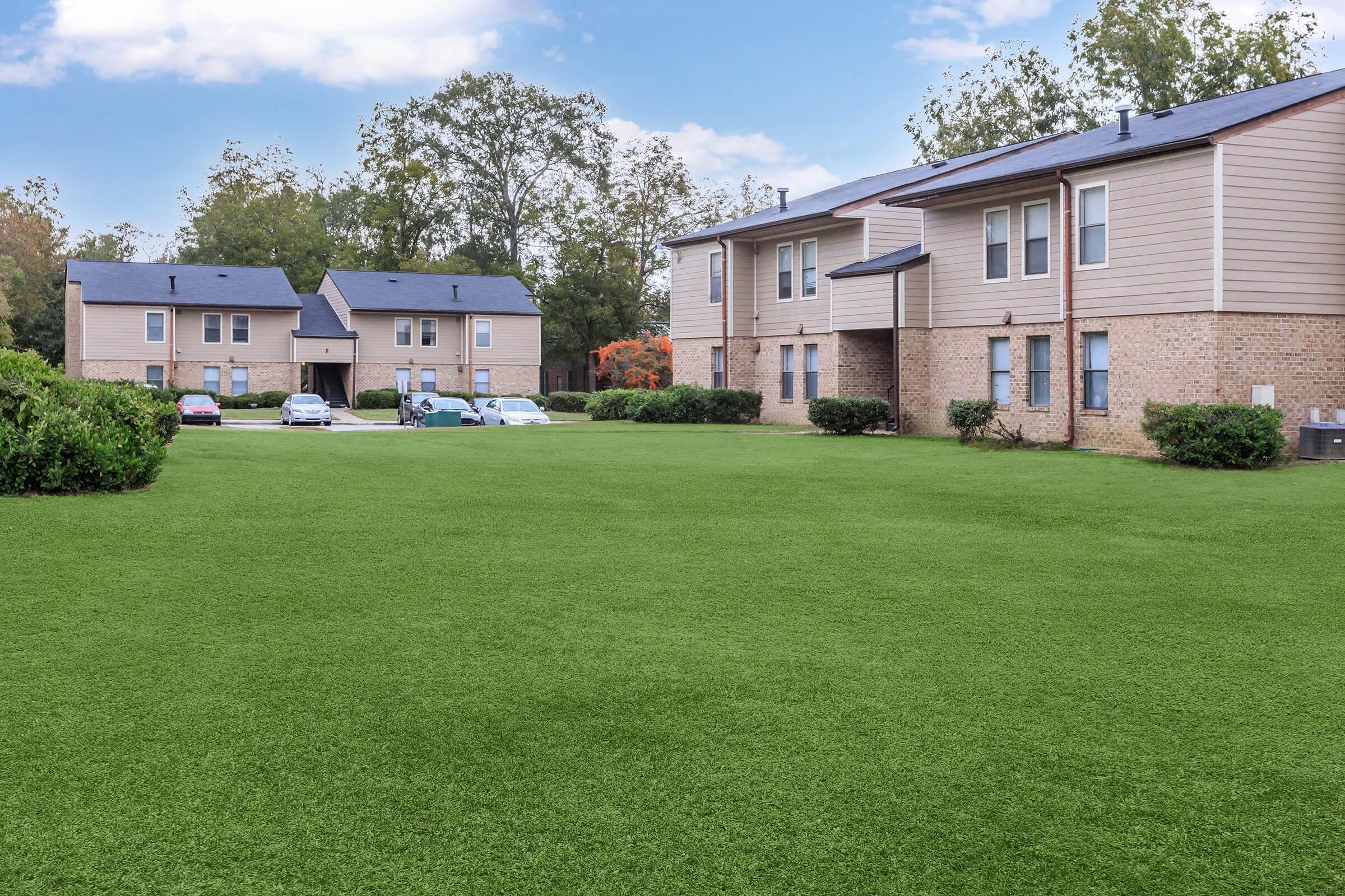A green, well-maintained lawn in front of two residential buildings. The buildings are light-colored with multiple windows and solar panels on the roofs. Several cars are parked nearby, surrounded by neatly trimmed bushes and trees in the background. The sky is clear with a few clouds.
