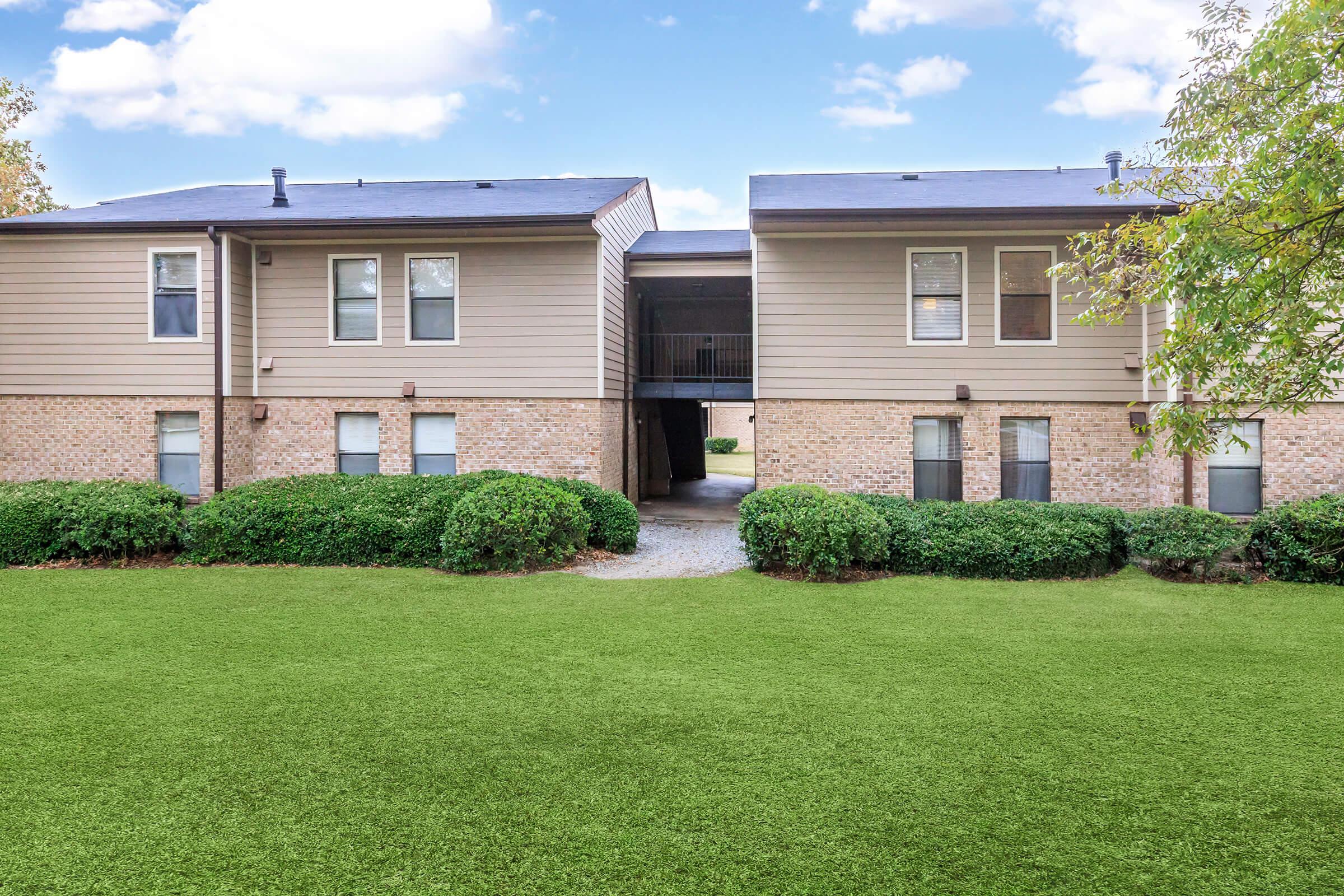 Two-story apartment buildings with beige siding and brick accents, separated by a grassy lawn. The buildings feature several windows and a walkway leading between them. Soft clouds are visible in the blue sky overhead, creating a tranquil atmosphere.
