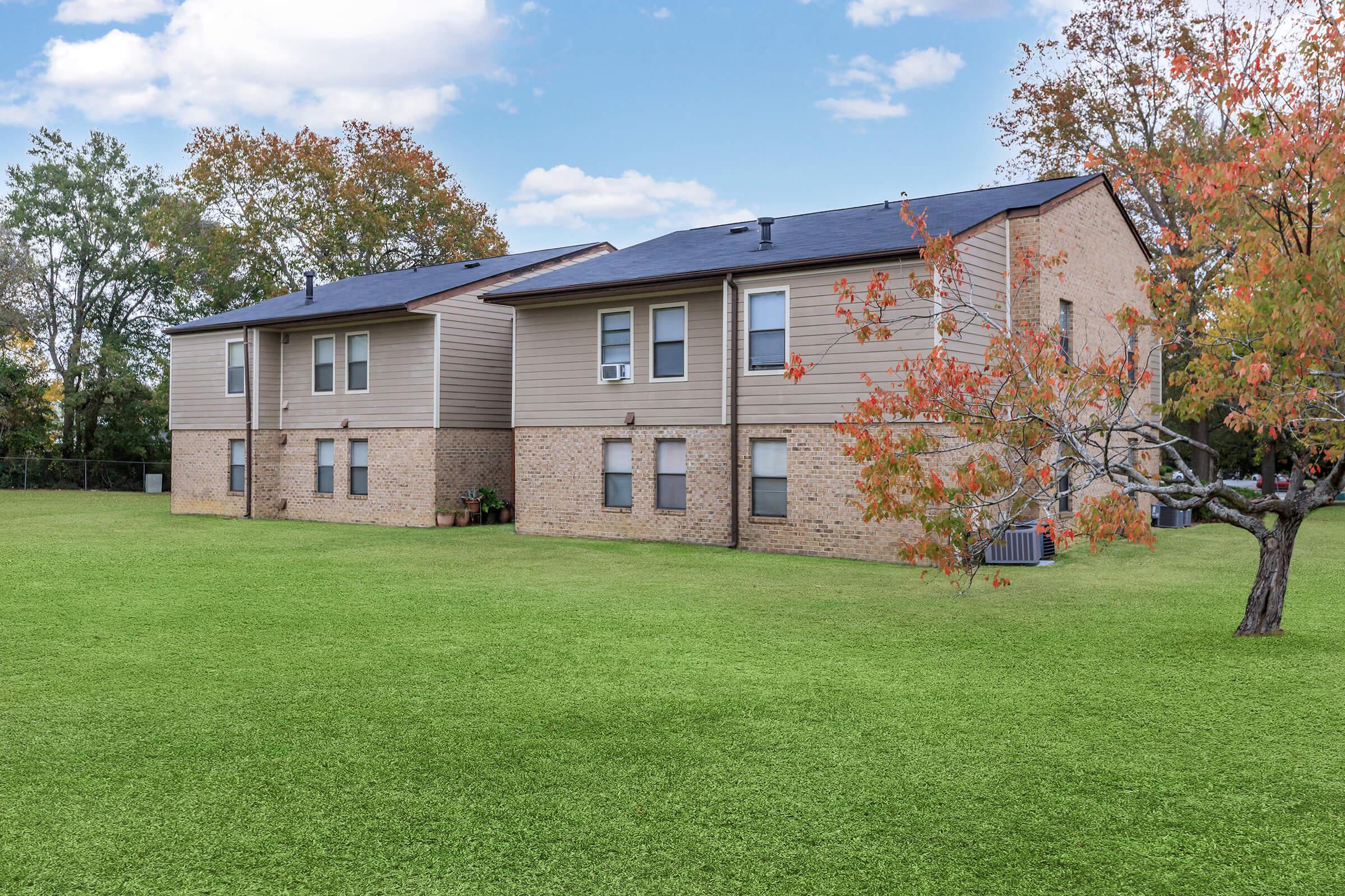Two suburban apartment buildings with light brown siding and small windows, surrounded by a well-maintained green lawn. A tree with autumn-colored leaves is in the foreground, adding to the serene atmosphere under a clear blue sky with scattered clouds.
