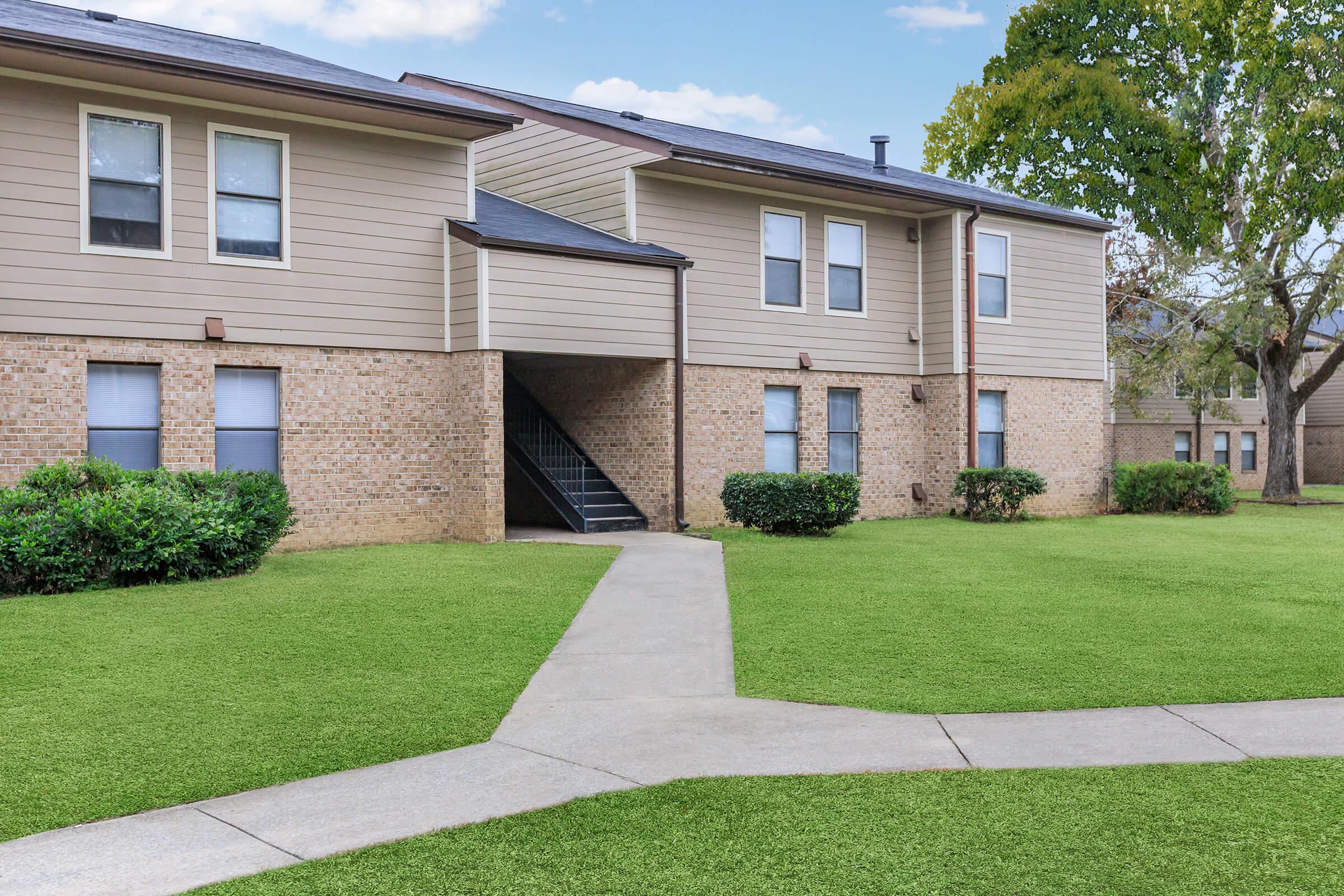 Exterior view of a two-story apartment building with a combination of brick and light-colored siding. The building features several windows and a staircase leading up to the second floor. Lush green lawns and neatly trimmed shrubs surround the building, adding to the aesthetic.