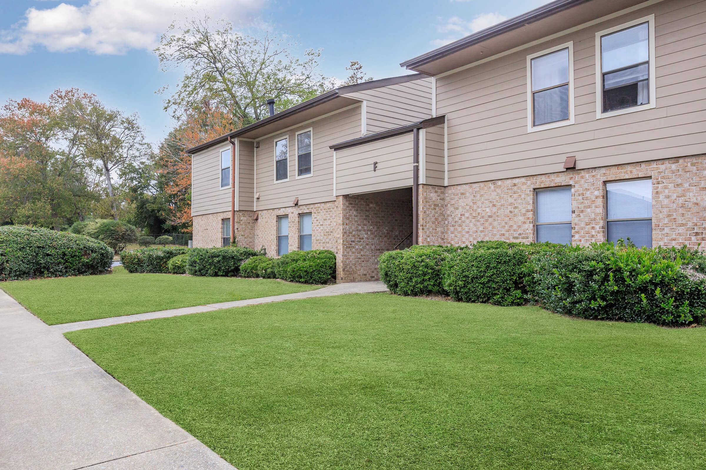 A well-maintained residential building with a brick and siding exterior, surrounded by neatly trimmed hedges and lush green grass. A clear walkway leads to the entrance, and trees with autumn foliage are visible in the background, creating a serene and inviting atmosphere.