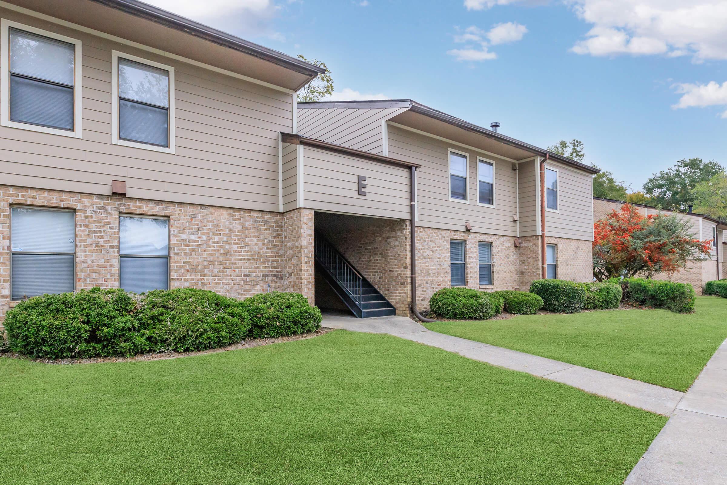 A multi-unit residential building featuring a combination of brick and siding exterior. The structure has two levels, with windows and a staircase leading to upper units. Well-maintained landscaping includes green grass and shrubbery lining the pathway. The sky is clear with a few clouds.
