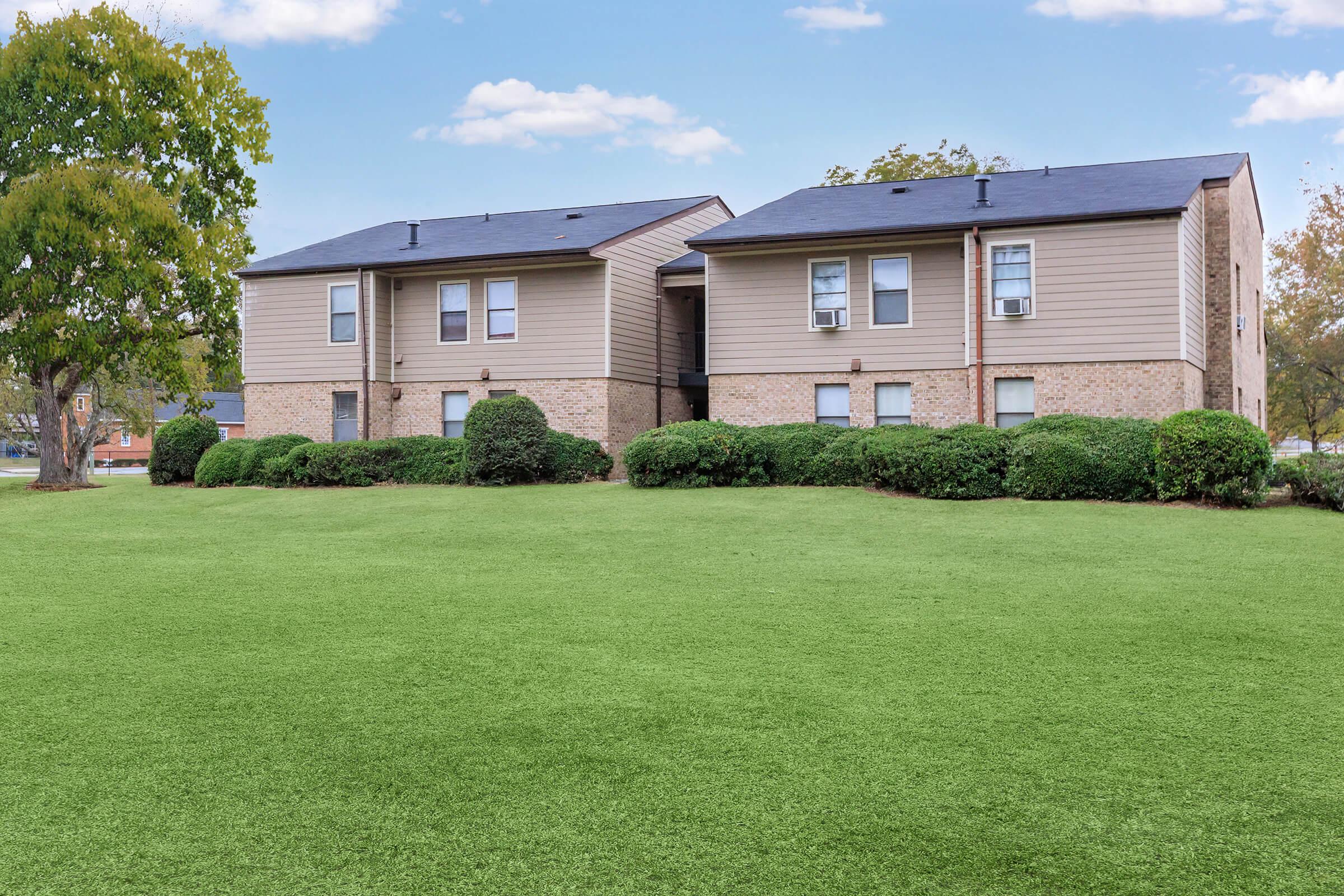Two residential buildings with a mix of brick and siding exteriors are surrounded by well-maintained green lawns and shrubs. The sky is clear with a few clouds, creating a pleasant outdoor atmosphere.