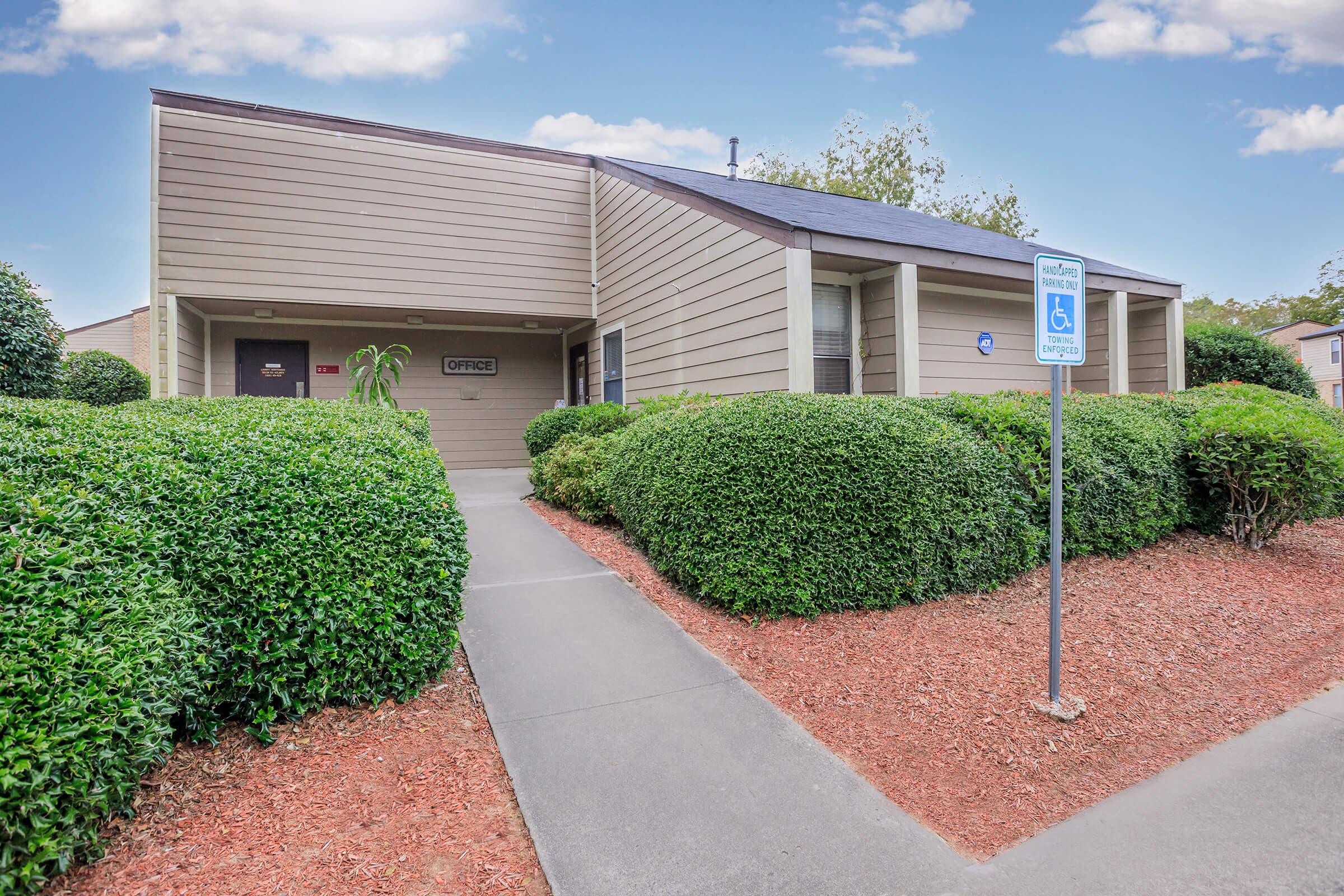 A single-story building with a sloped roof and light brown exterior. The entrance features two doors labeled "Office" and is flanked by neatly trimmed green hedges. A pathway leads up to the entrance, and there's a handicapped parking sign visible nearby. The sky is clear with a few clouds.