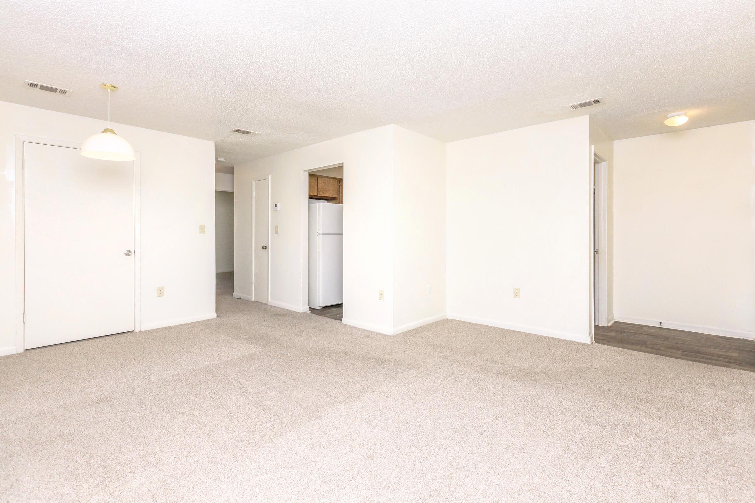 Spacious, empty living area with beige carpet and white walls. A hallway leads to a kitchen area featuring a white refrigerator. The ceiling has a simple light fixture, and doors on the left lead to an entryway and other rooms. Bright and airy atmosphere with a clean, uncluttered look.