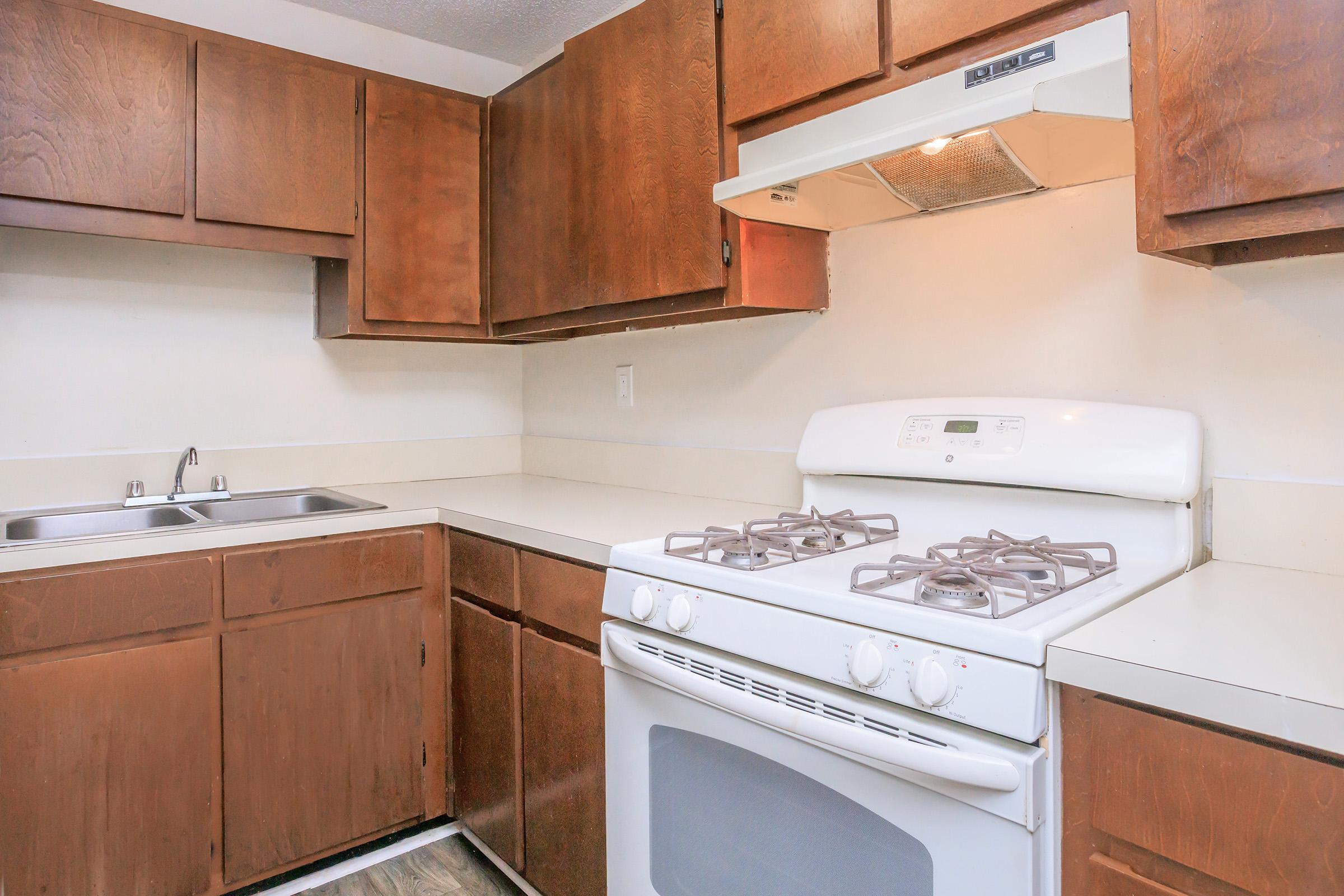 A kitchen with wooden cabinets, a white gas stove with an oven, and a double sink. The countertops are light-colored, and the kitchen has a neutral color scheme. A range hood is above the stove, and there's ample storage space in the cabinets.