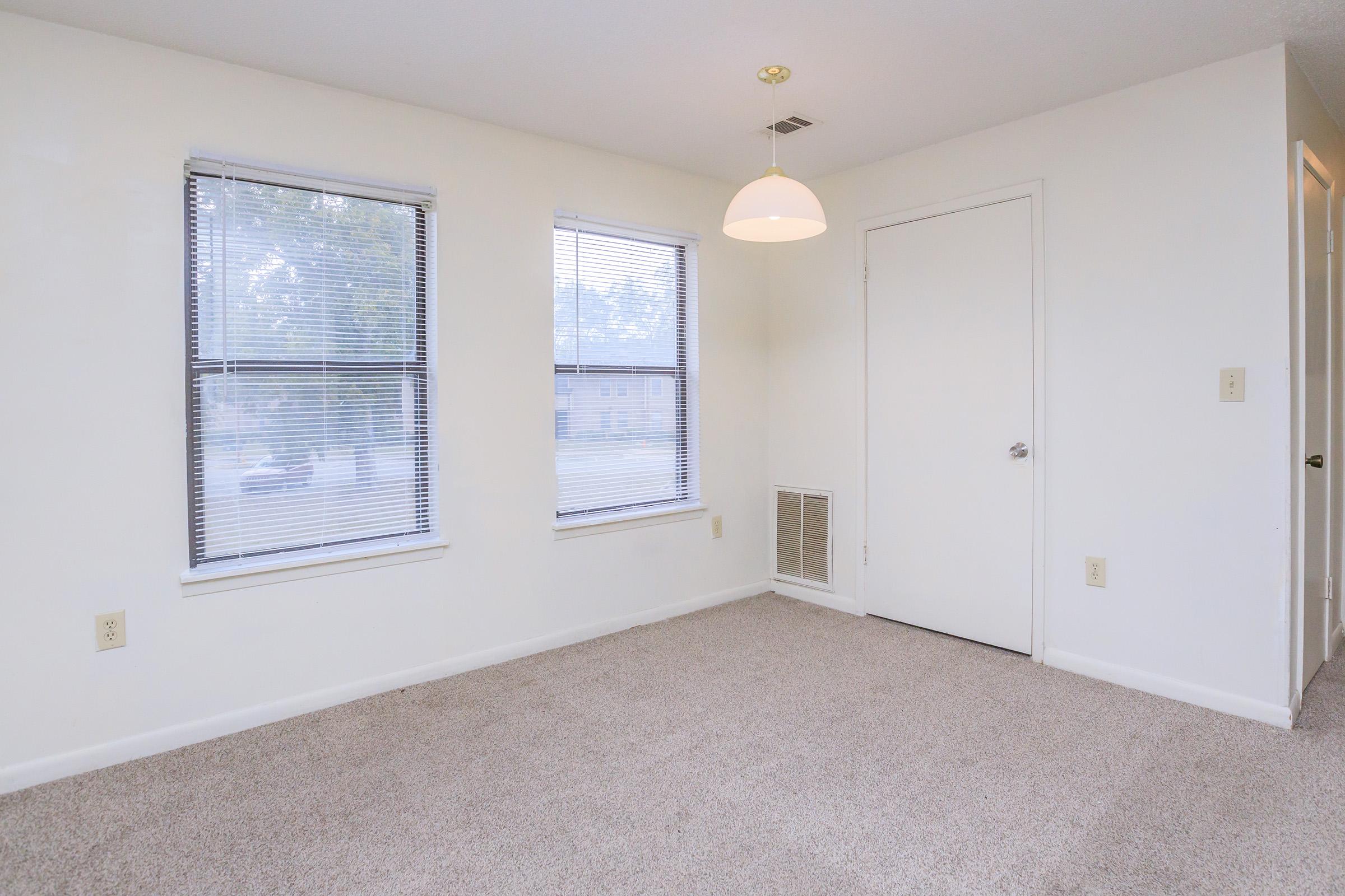 A bright, empty room with light beige carpet, two windows with blinds, and a single pendant light hanging from the ceiling. The walls are painted white, and there is a closed door on one side. A vent is positioned near the floor, and there are no furnishings or decorations present.