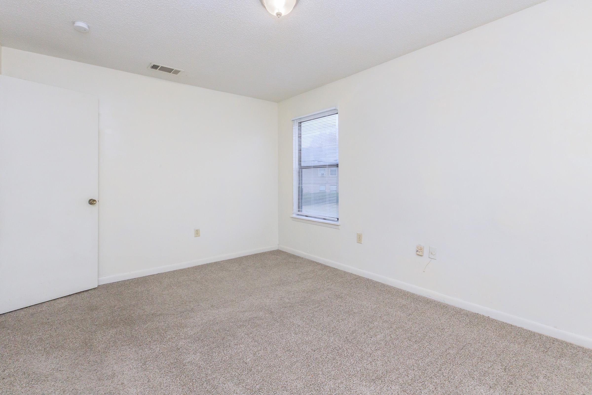 A spacious, empty room with light-colored walls and a small window. The floor is covered in beige carpet, and there is a closed white door on the left side. Natural light enters through the window, highlighting the simplicity of the space.